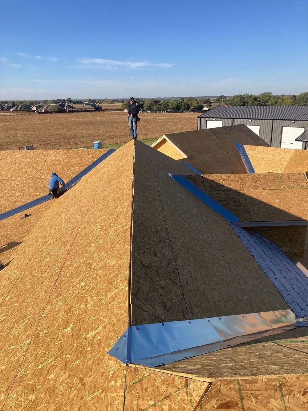 Construction workers on a roof, installing roofing materials. Blue sky, open field visible in the background.