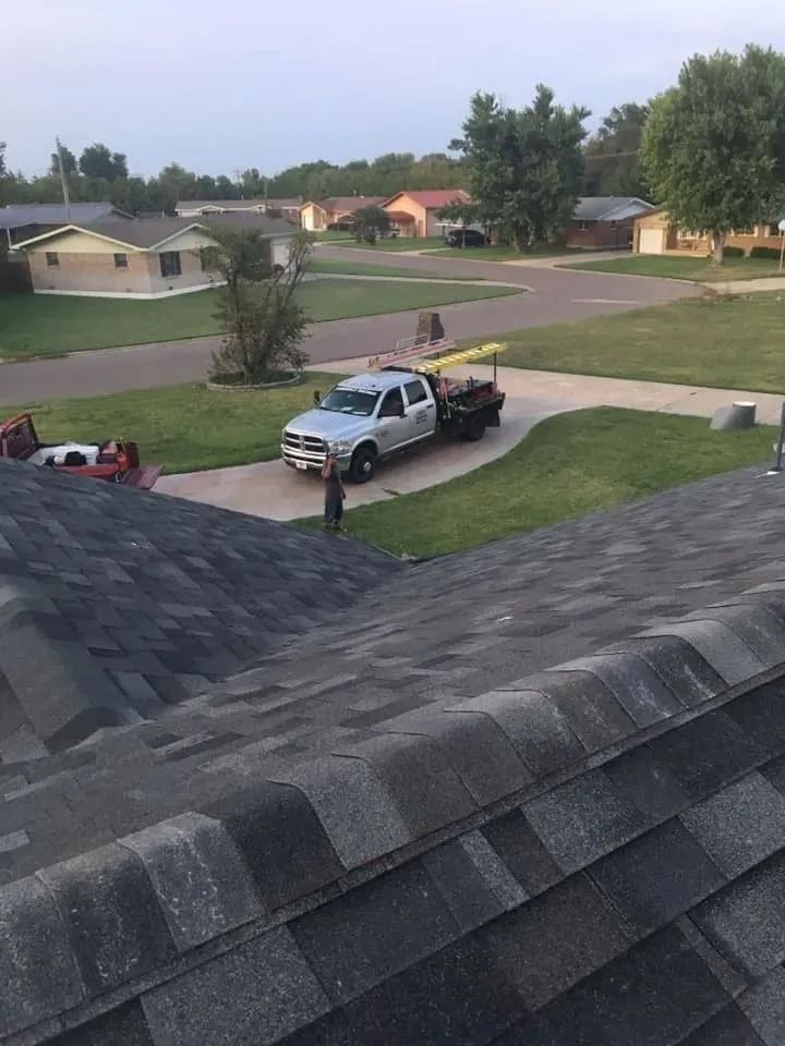 View from a dark shingled roof, looking at a truck with a trailer and a person on a lawn in a residential area.