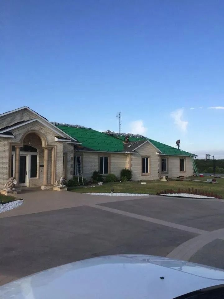 A house with roof damage, green underlayment exposed.  Blue sky and driveway visible.