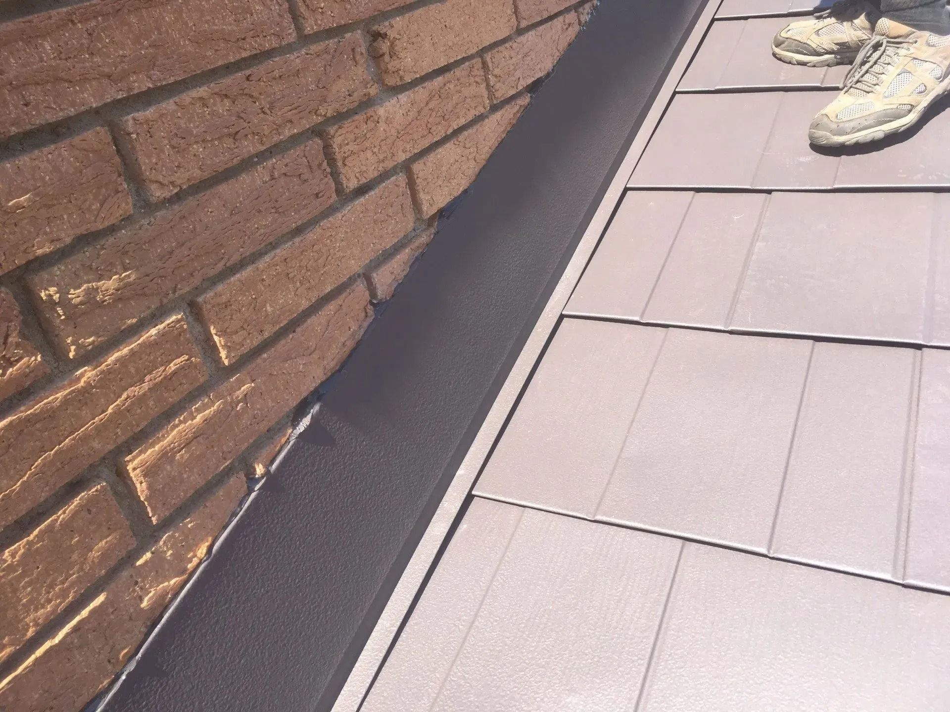 Close-up of a brick wall and roof tiles with a black flashing strip between them. A person's feet are visible.