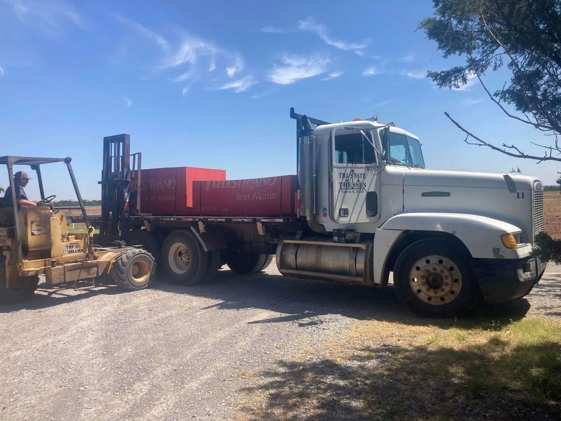 Forklift unloading red blocks from a white semi-truck on a gravel road, sunny day.