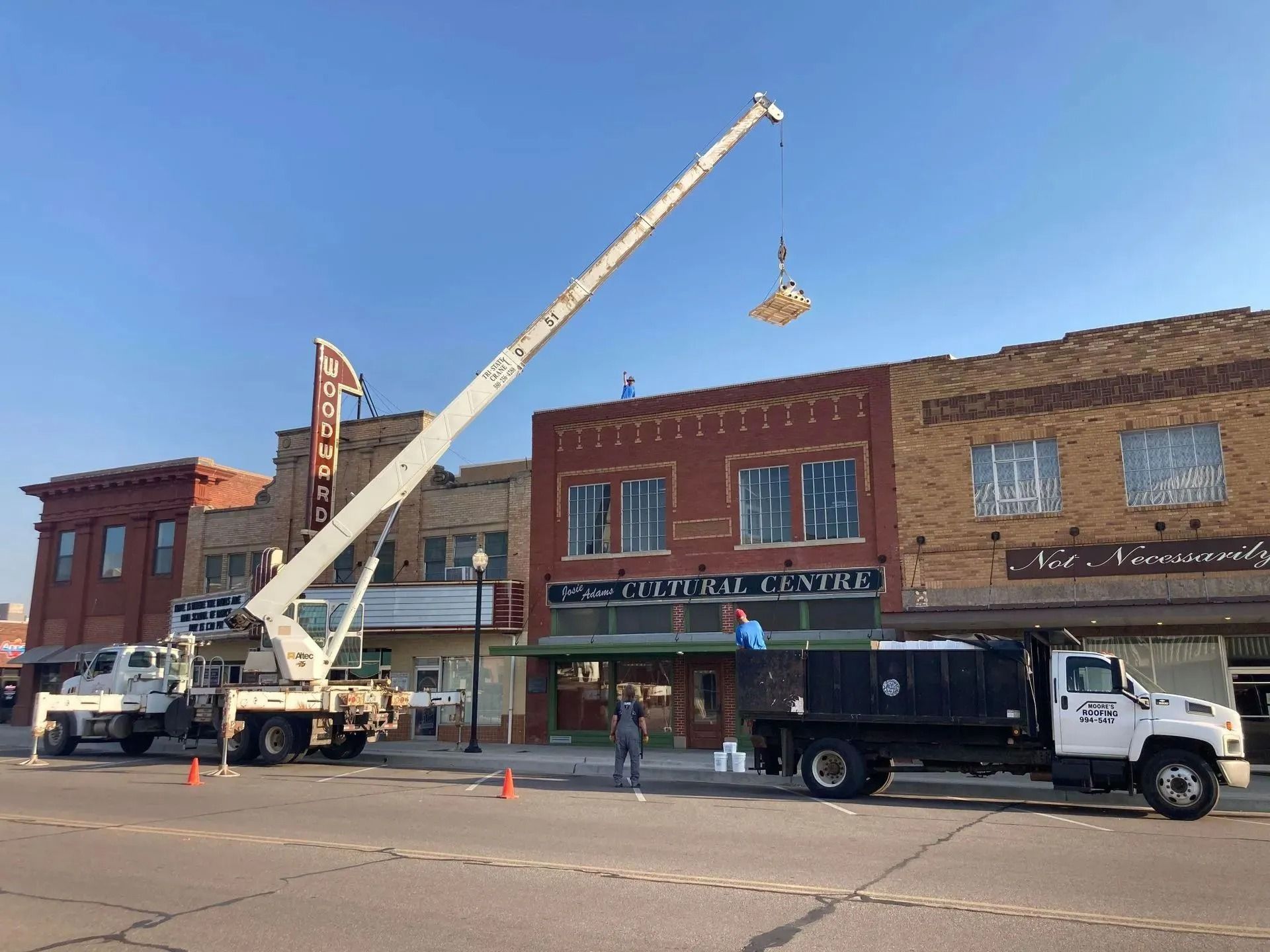 A crane lifts an object above a downtown street; worker stands nearby. Red brick buildings and trucks in view.