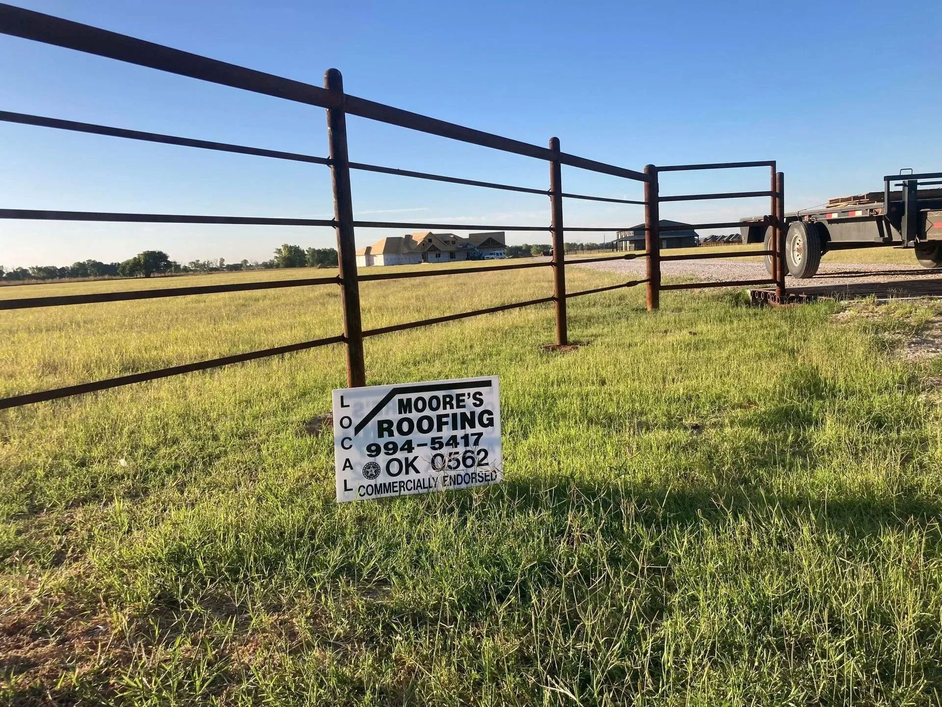 Sign for Moore's Roofing in front of a metal fence, grassy field, and a truck. Sky is blue.