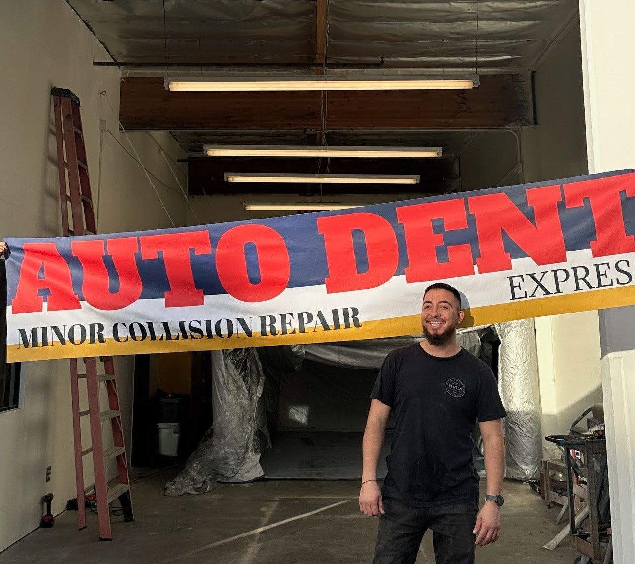 Man holding "Auto Dent Express" banner in an open garage space. Red, blue, and white banner, smiling.