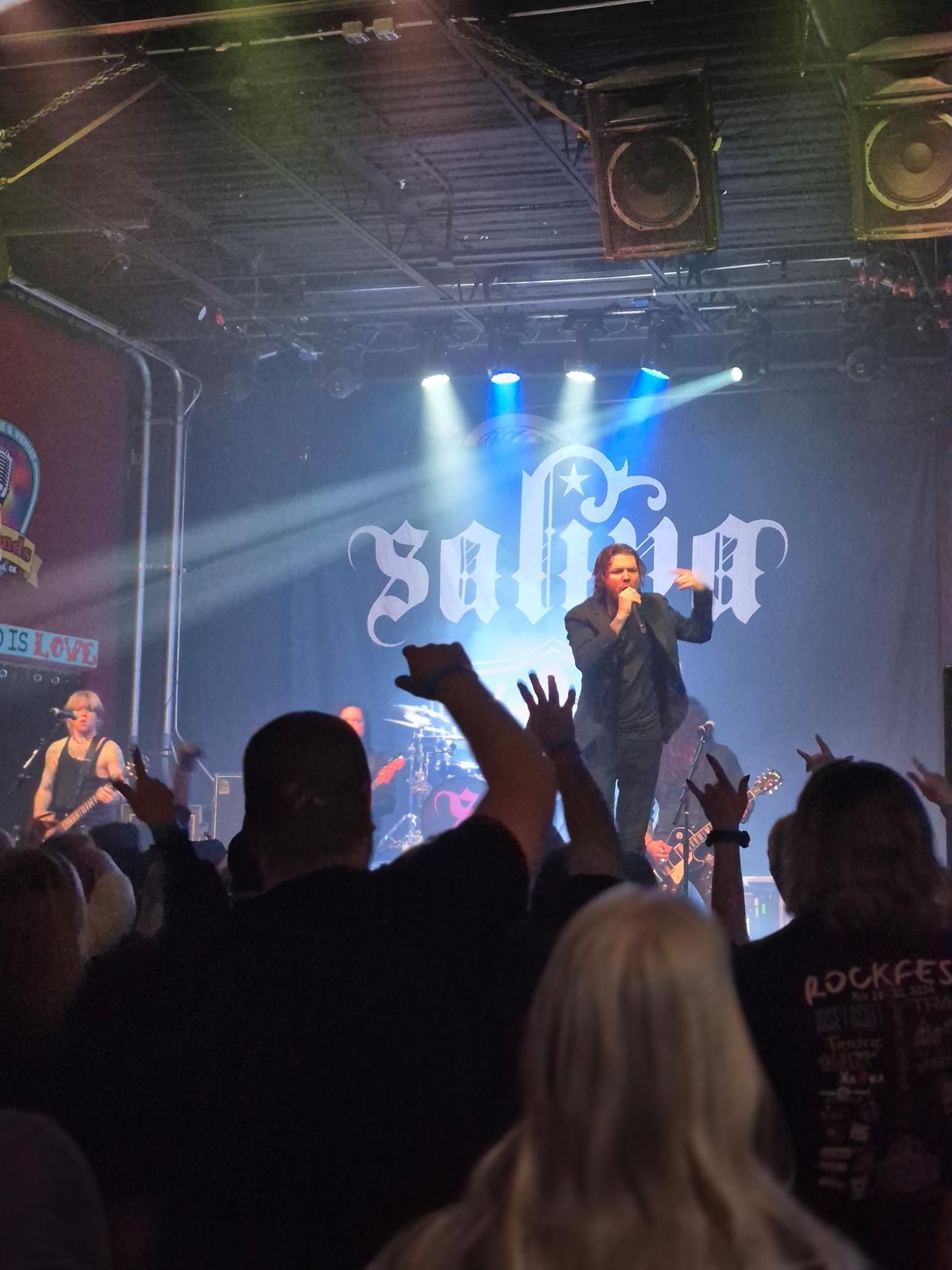 A band performing on stage in front of a crowd. Singer with an outstretched hand, logo Saliva.