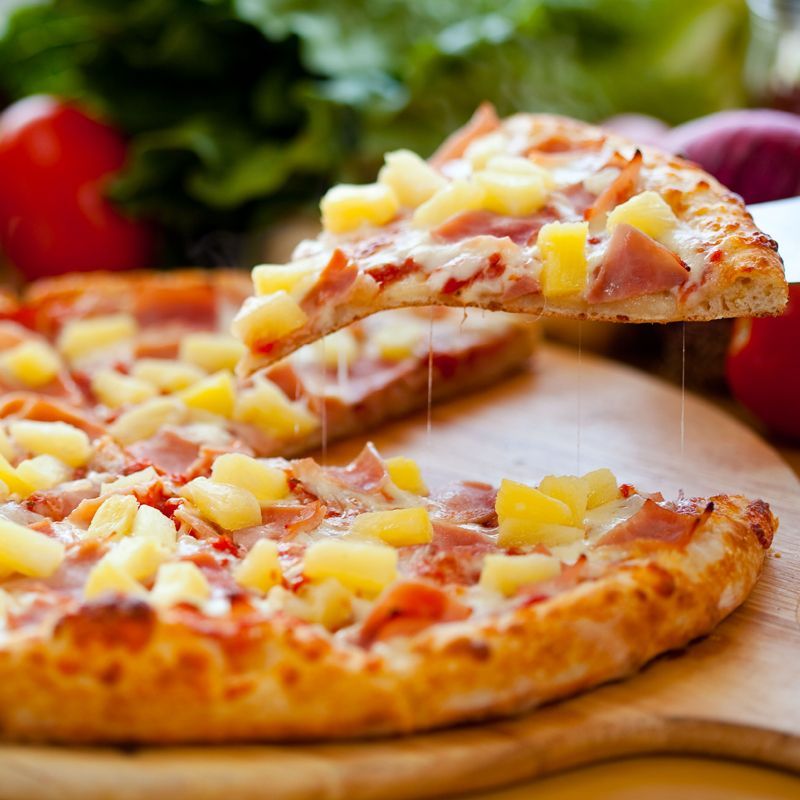 A pizza with pineapple and ham is being sliced on a wooden cutting board