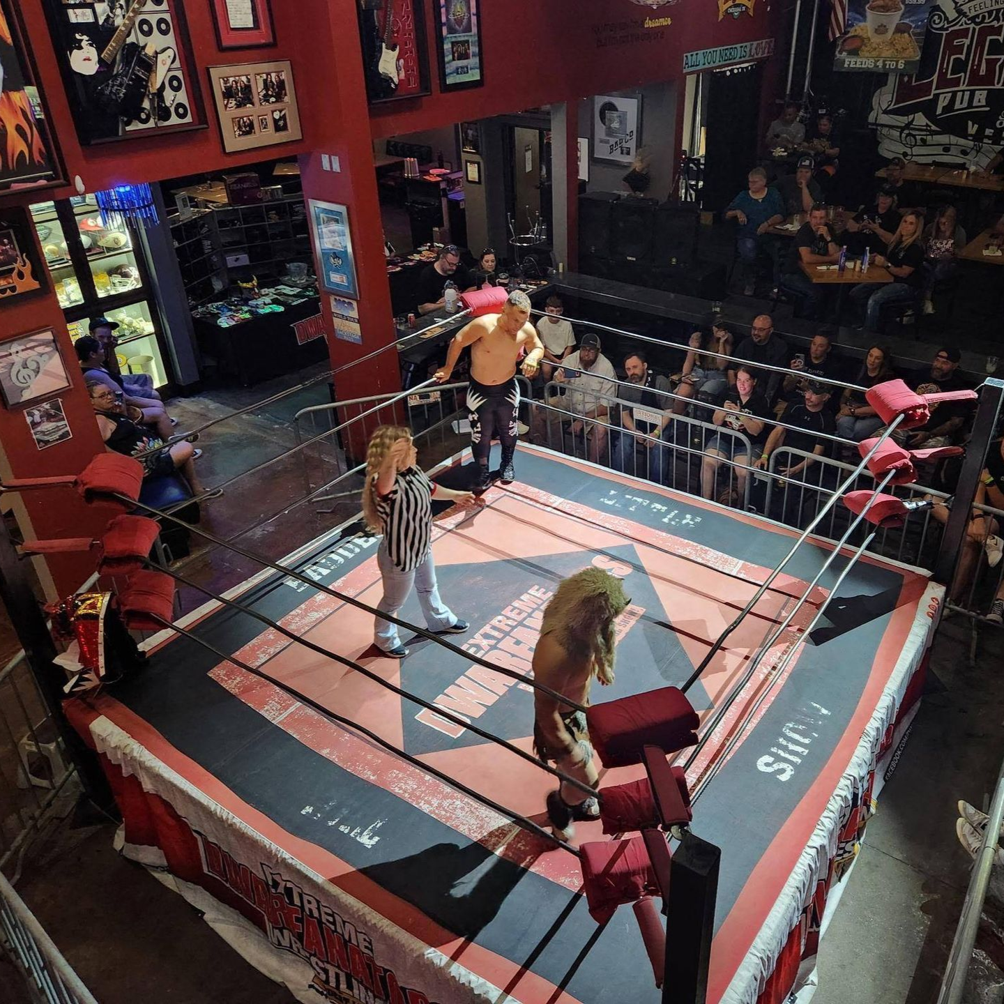 Wrestling match in a red-themed venue. Wrestlers and referee in a ring, audience watching from the sides.