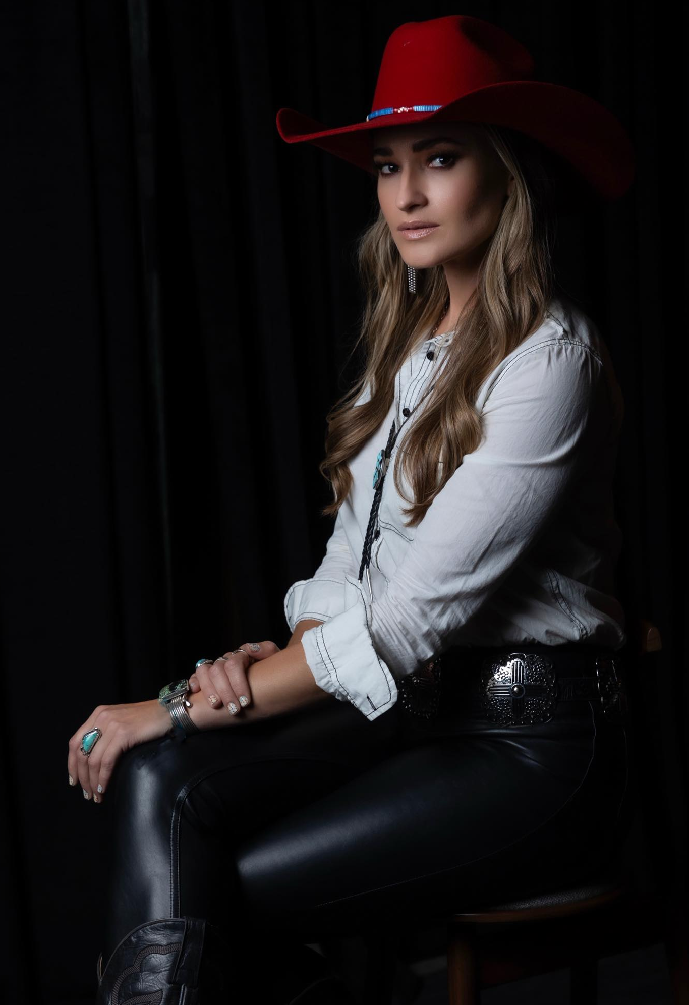 Woman in cowboy hat, white shirt, and black pants sits in a dimly lit setting.