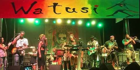 A group of people are playing instruments on a stage under a sign that says watusi.