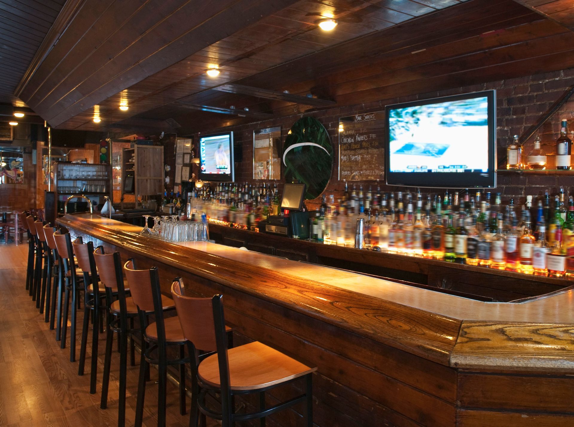 An empty wooden bar with a long counter, a row of stools, televisions on a brick wall, and shelves of liquor bottles.