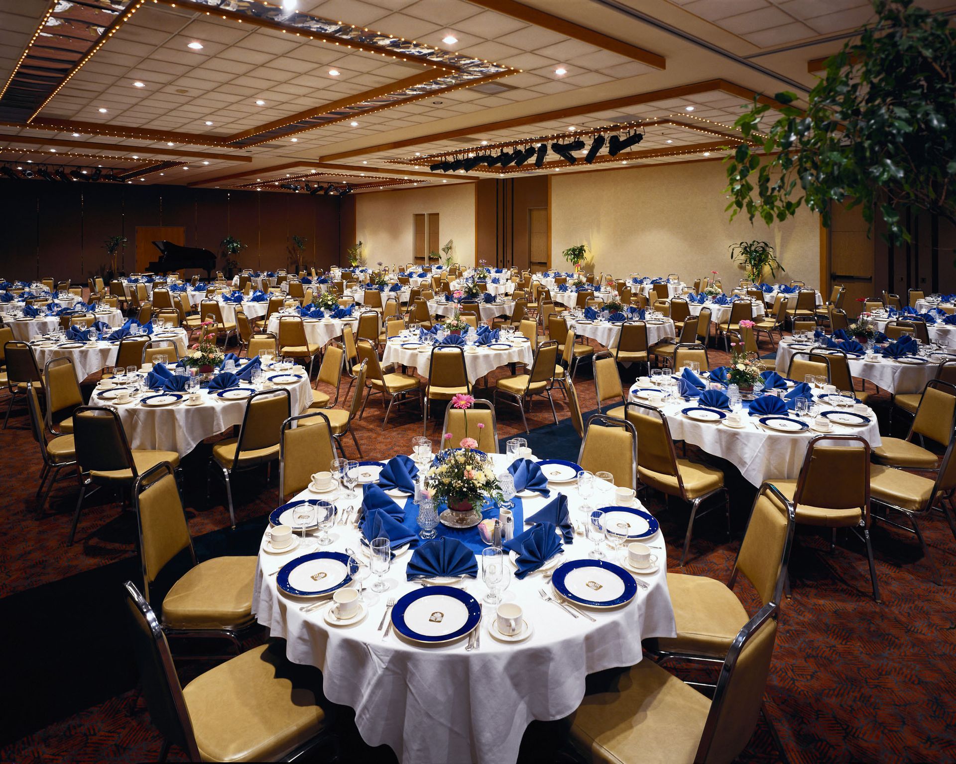 A banquet hall filled with round tables covered in white tablecloths, gold chairs, and blue napkins.