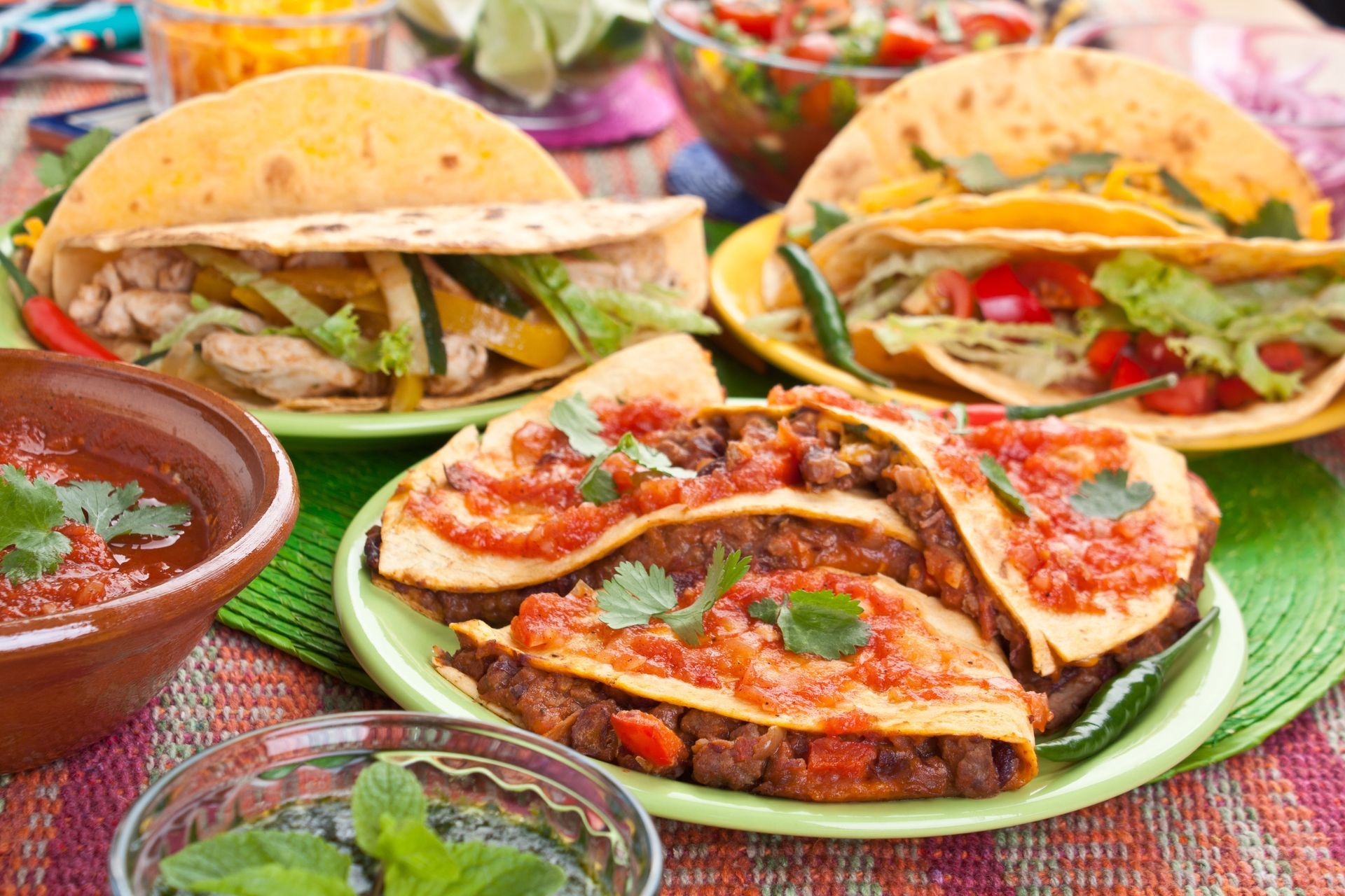 A spread of tacos and quesadillas topped with salsa and herbs, with bowls of salsa and cilantro on a colorful tablecloth.