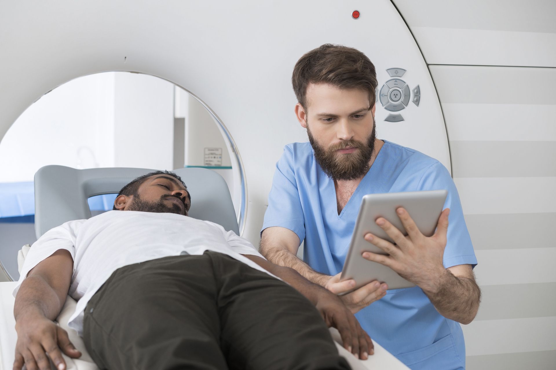 Man lying on a medical scanner as a healthcare worker in blue scrubs reviews a tablet.
