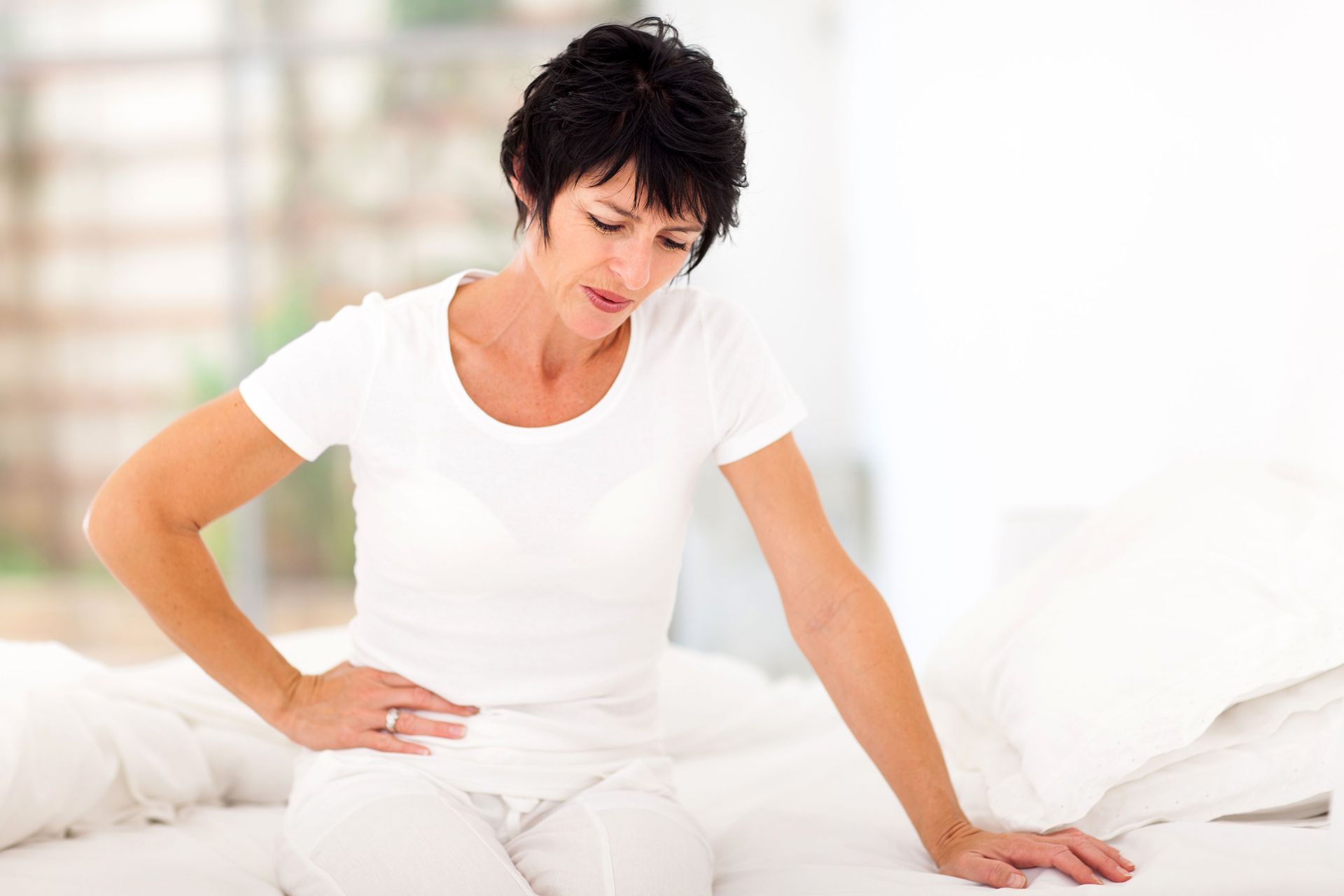 Woman clutching her side on a bed, showing discomfort, in a bright bedroom setting.