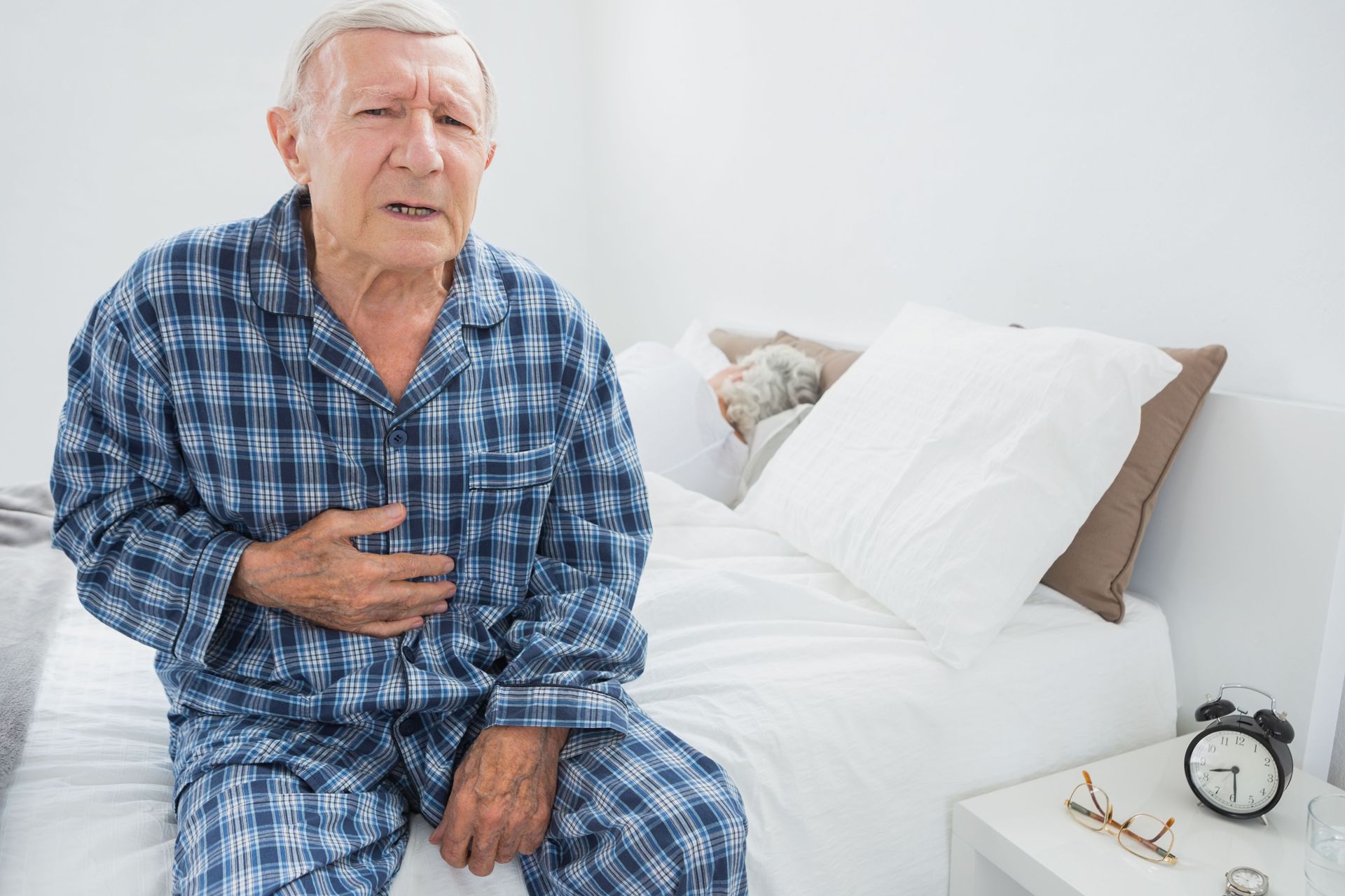 Man in blue pajamas clutches stomach, seated on a bed, appearing in pain.