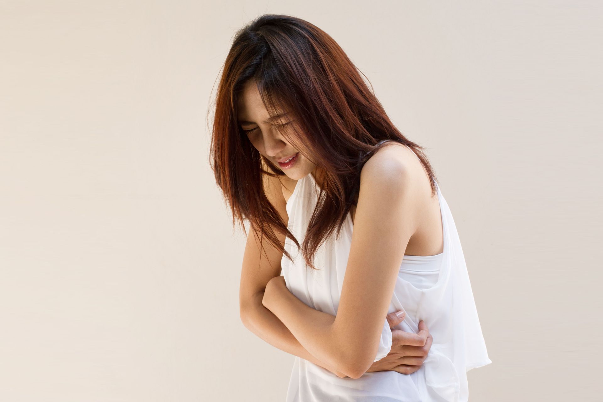 Woman in white top, holding stomach, appearing to be in pain, against a tan background.