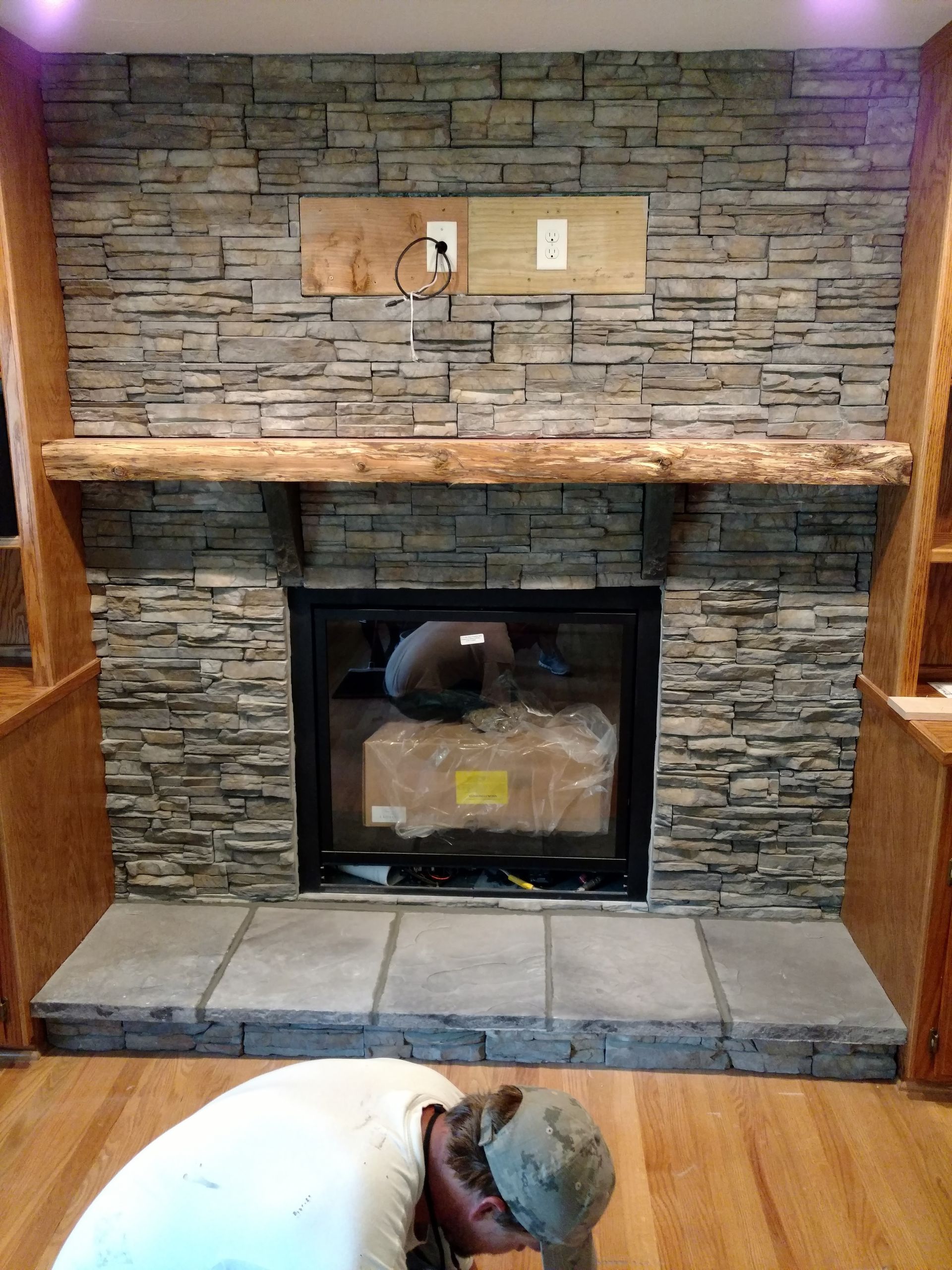 Stone fireplace with wooden mantel and a person crouched on a hardwood floor in the foreground