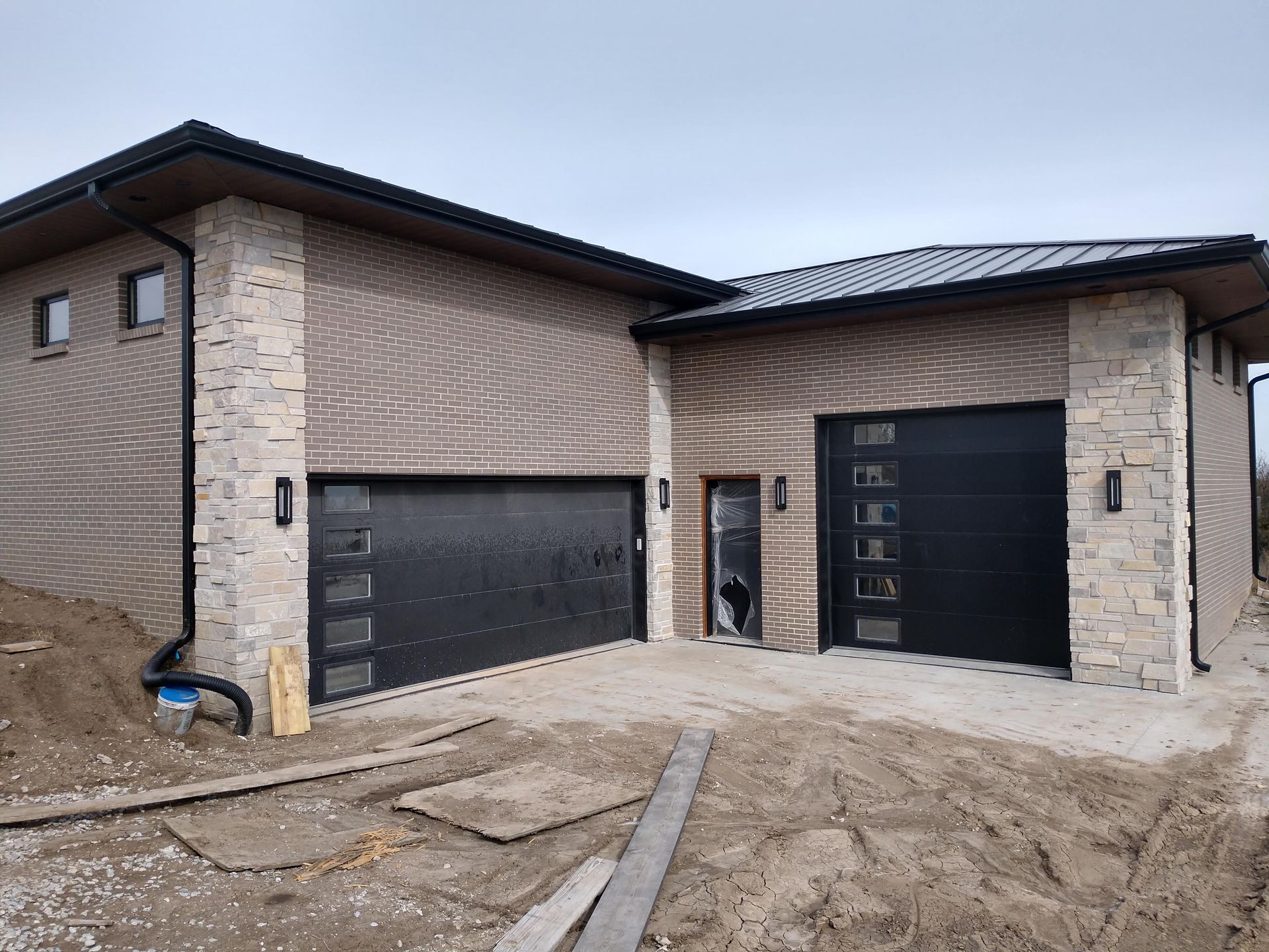 Modern two-story house with brick exterior, two black garage doors, and a muddy driveway under construction