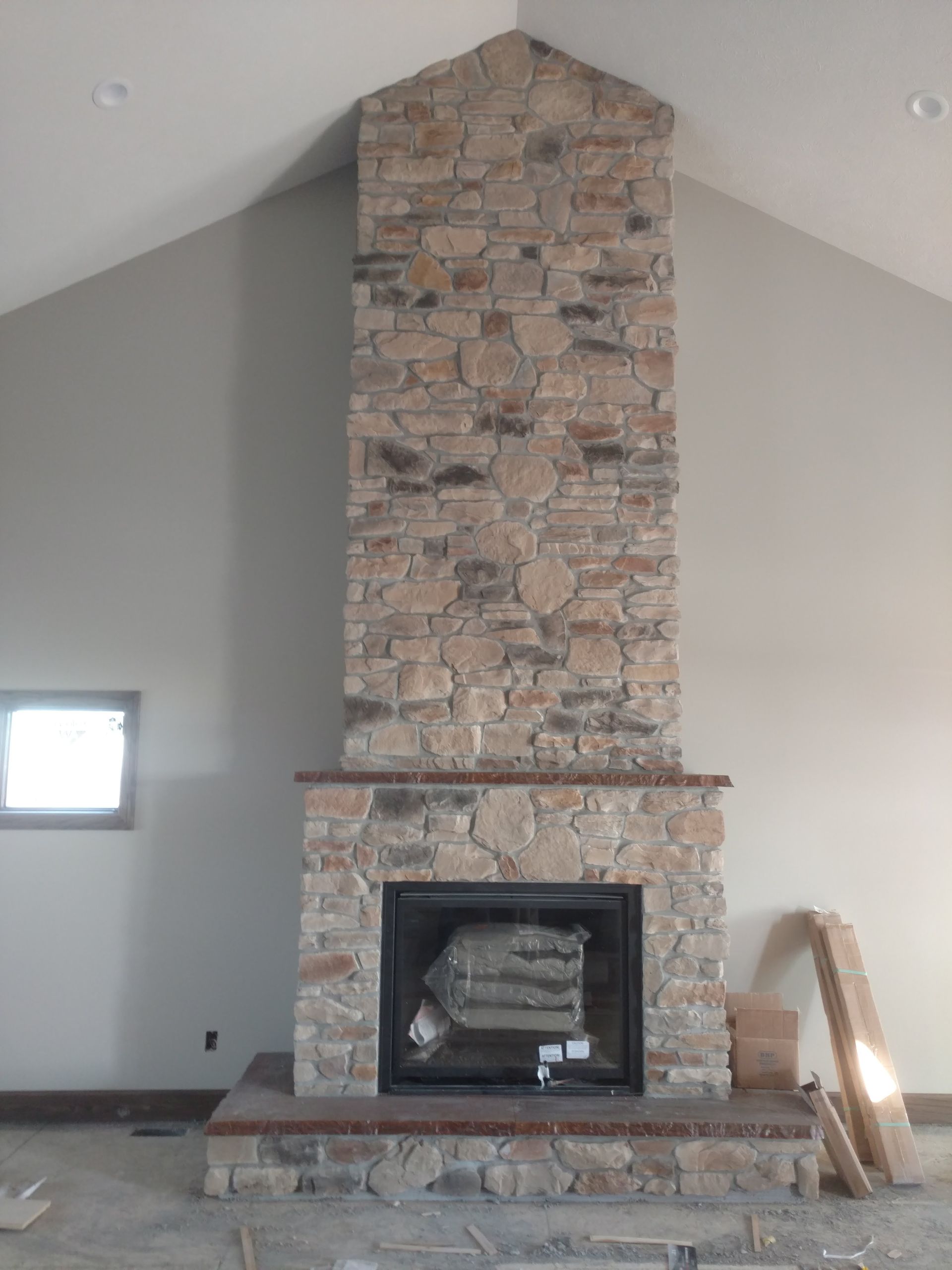 Stone fireplace with a tall chimney in a vaulted room, unfinished hearth with a small square window nearby