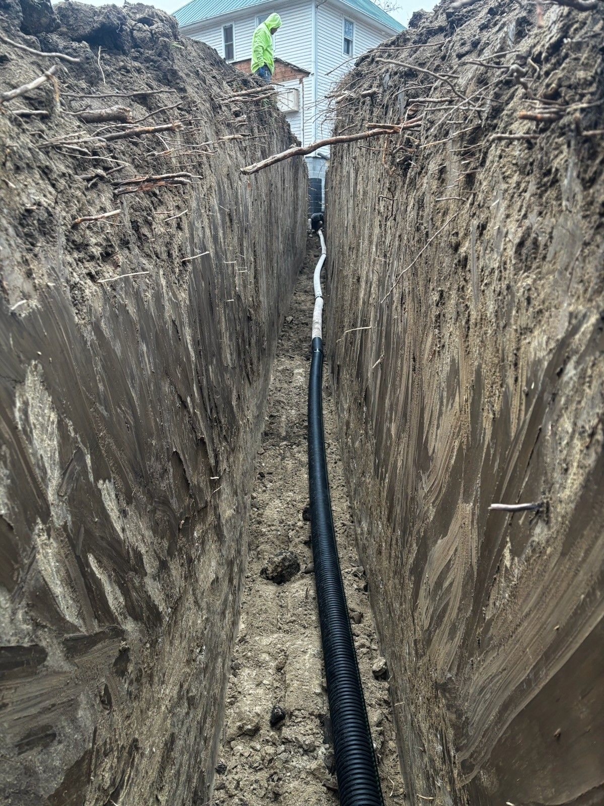 Deep trench with a black utility pipe laid along the bottom beside a house under construction
