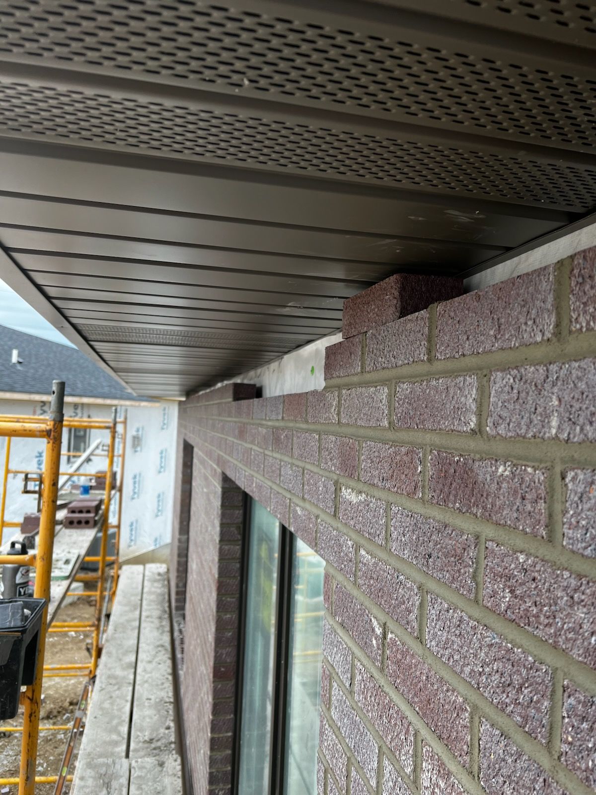 Exterior building wall with brick veneer and soffit, viewed from a narrow construction walkway with scaffolding.