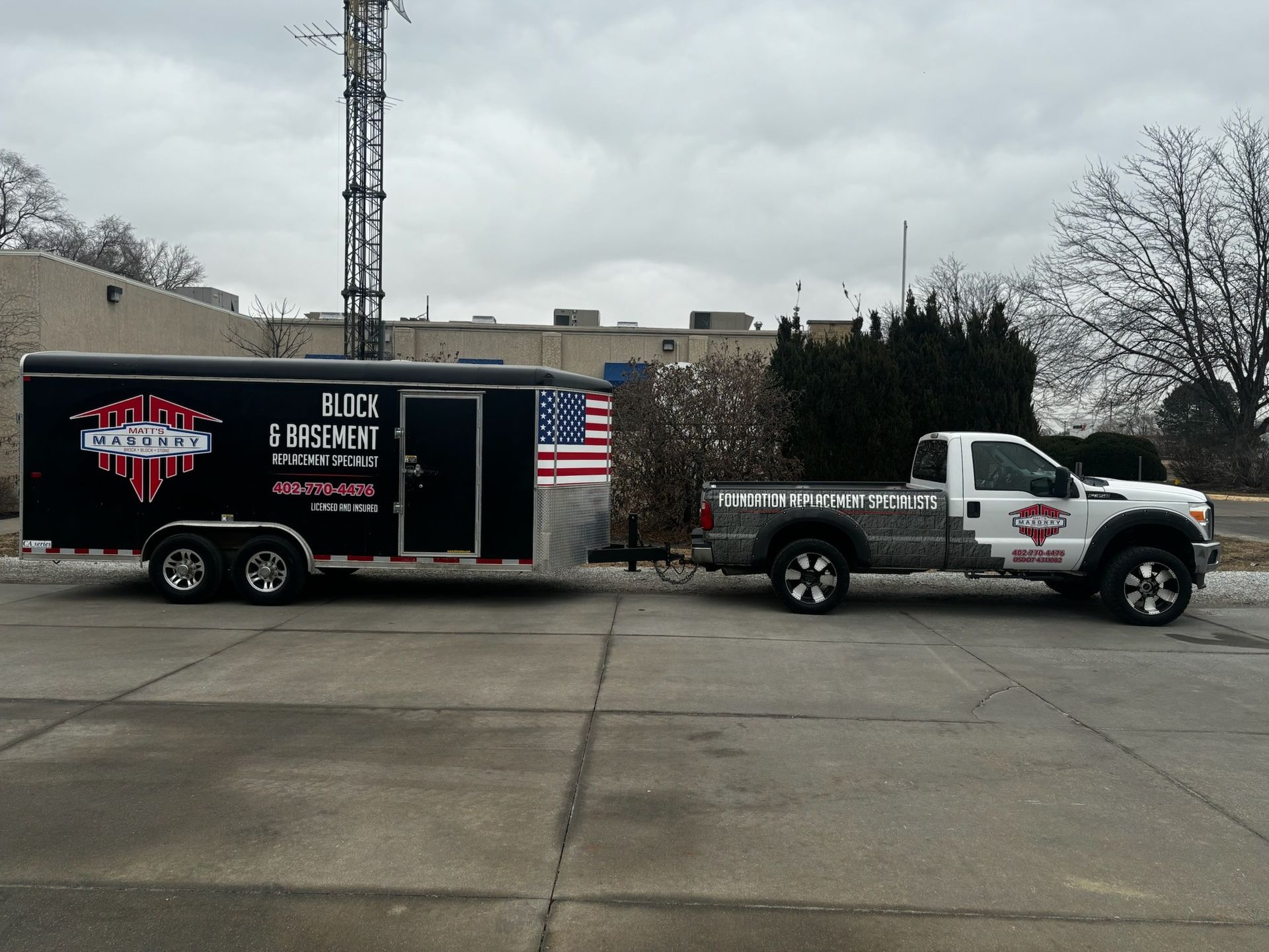 White pickup towing black-and-red trailer with American flag parked on wet pavement outdoors