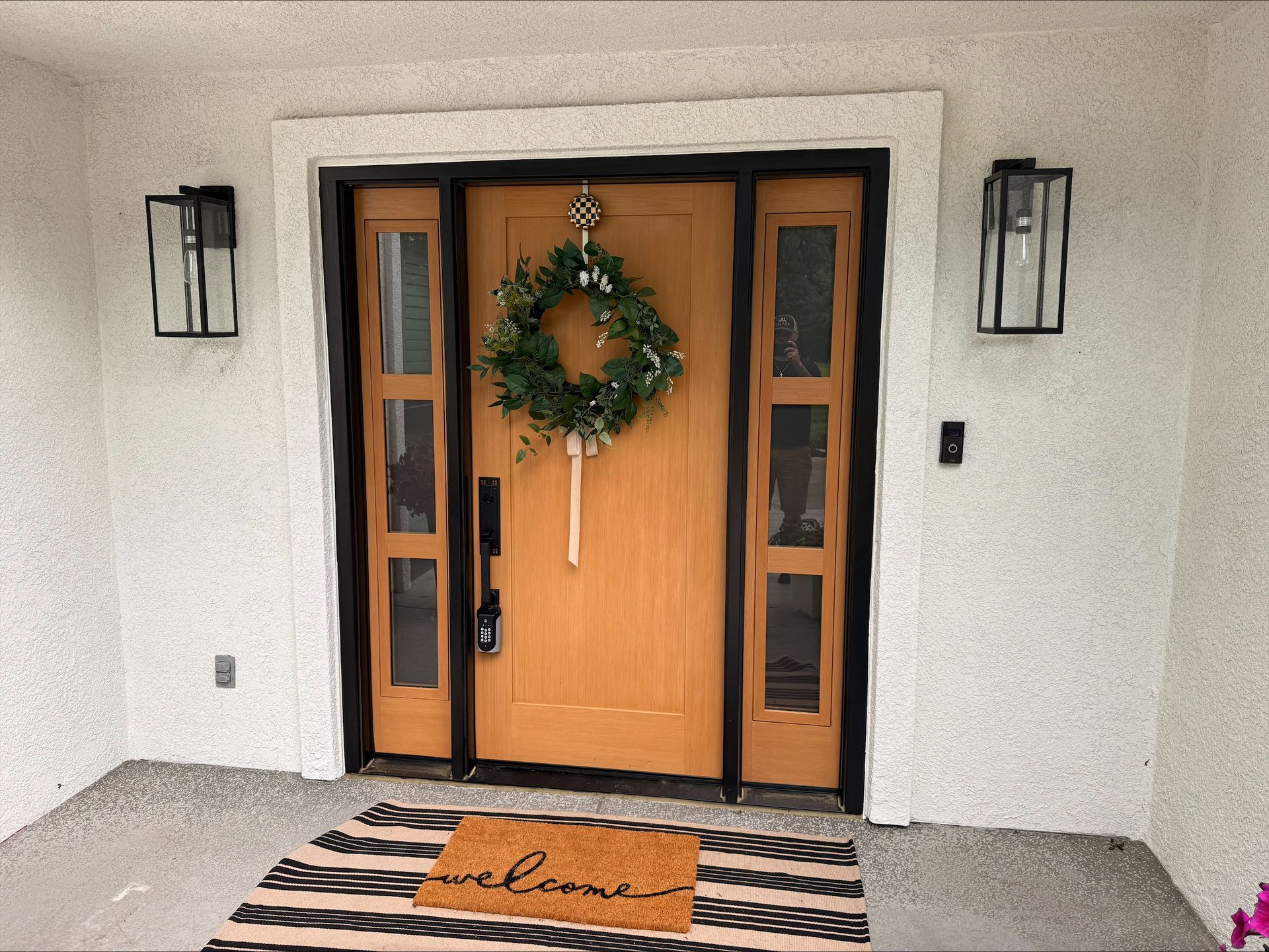 Light brown door with wreath and welcome mat, flanked by sidelights and black sconces.