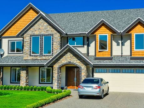 Two-story house with stone and beige siding, a gray roof, and a car in the driveway on a sunny day