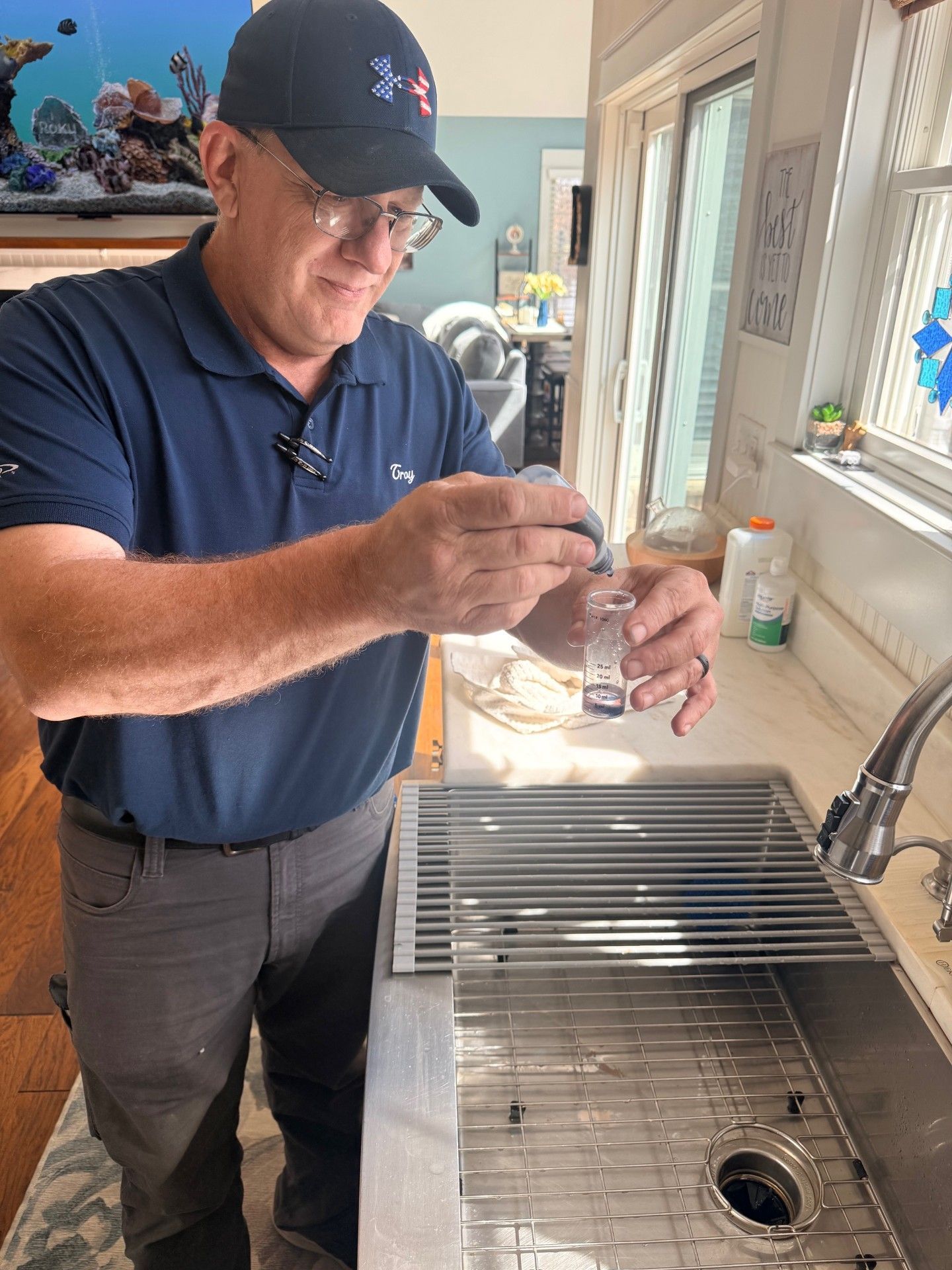 A person in a blue shirt and baseball cap adds a drop of liquid from a dropper bottle into a small vial above a kitchen sink.