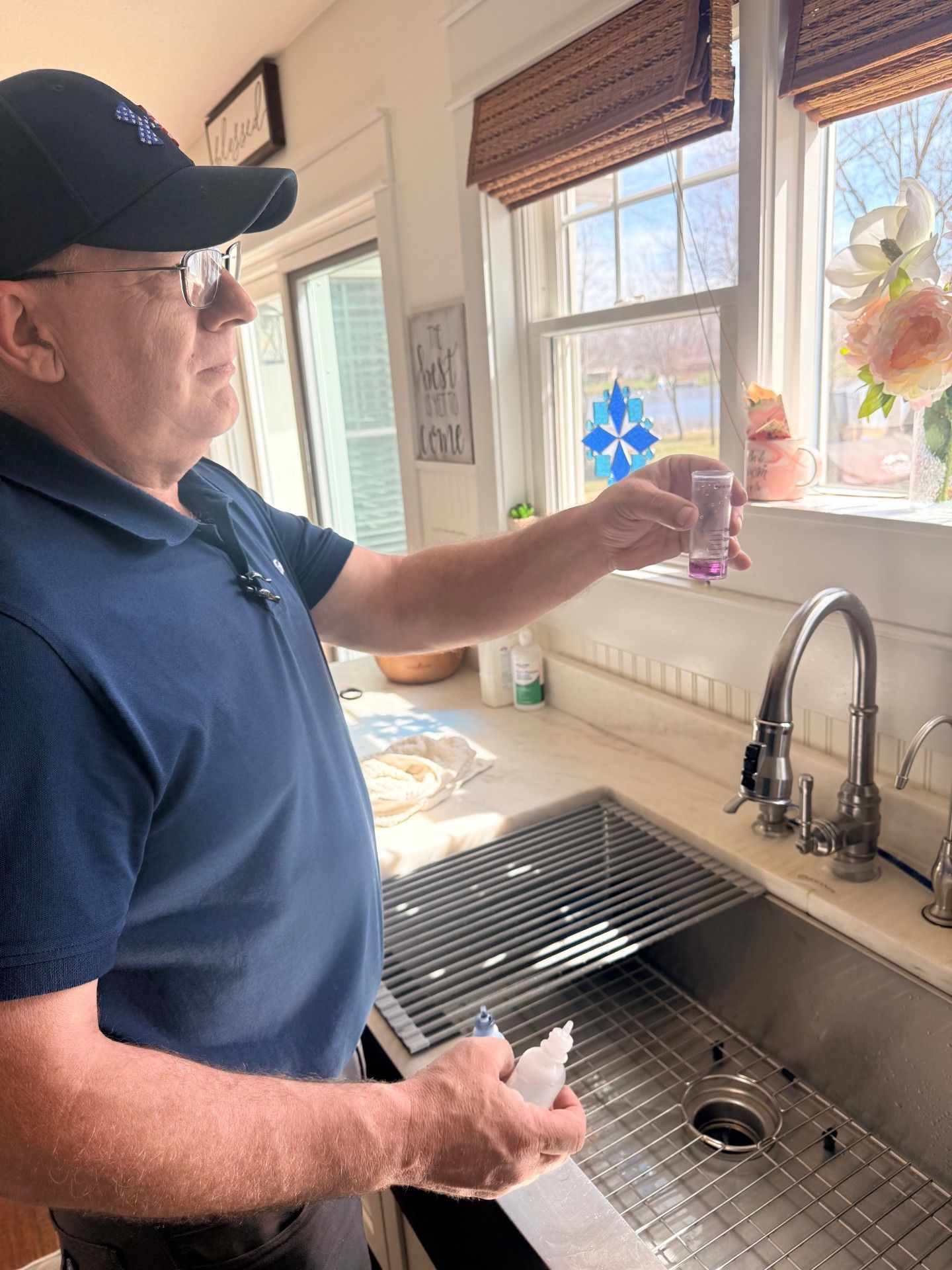 A person in a blue polo shirt and cap holds a small vial with purple liquid while standing in a kitchen by the sink.