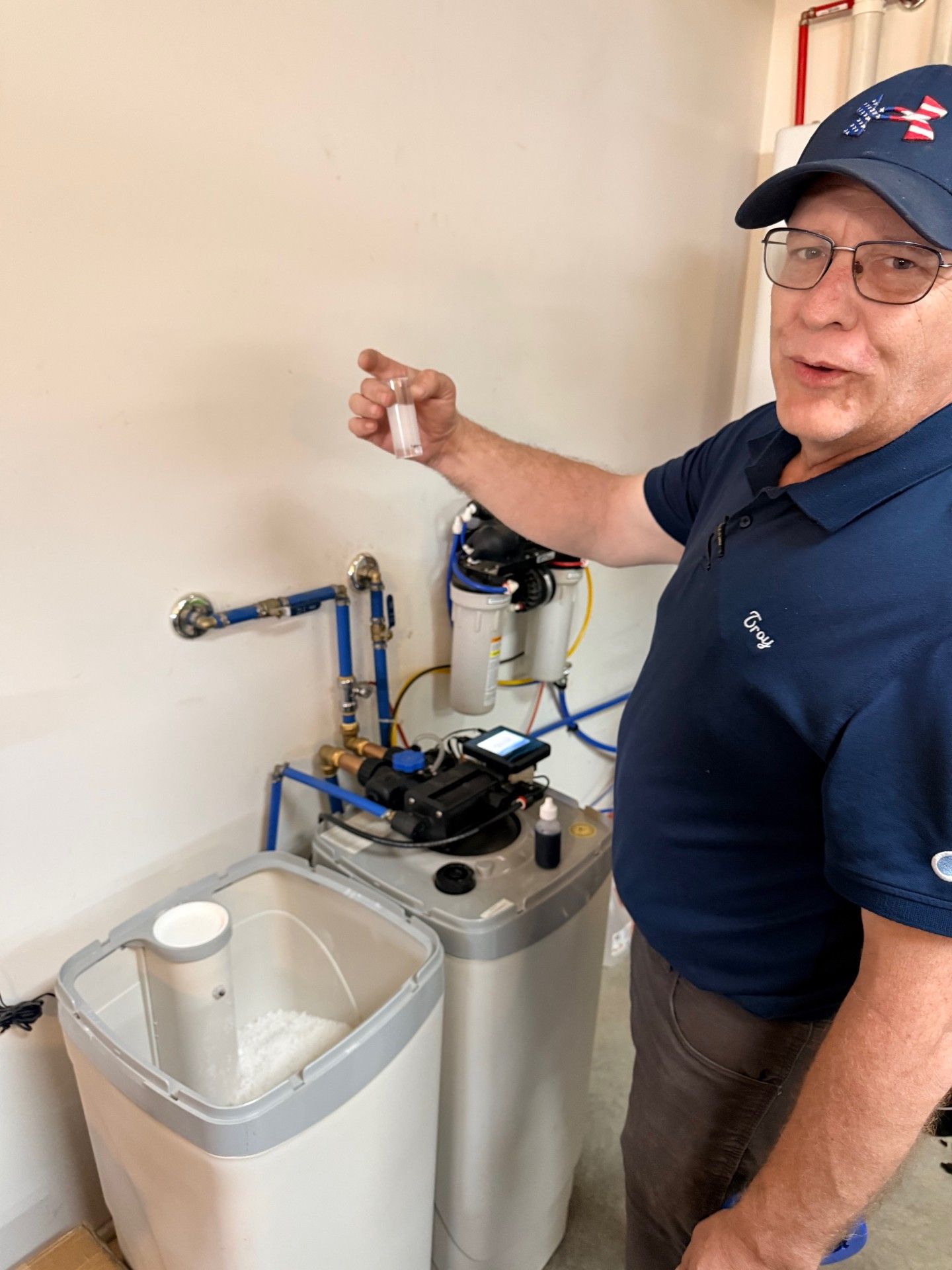A person in a blue polo and cap holding a small vial while standing beside a home water filtration and softening system.