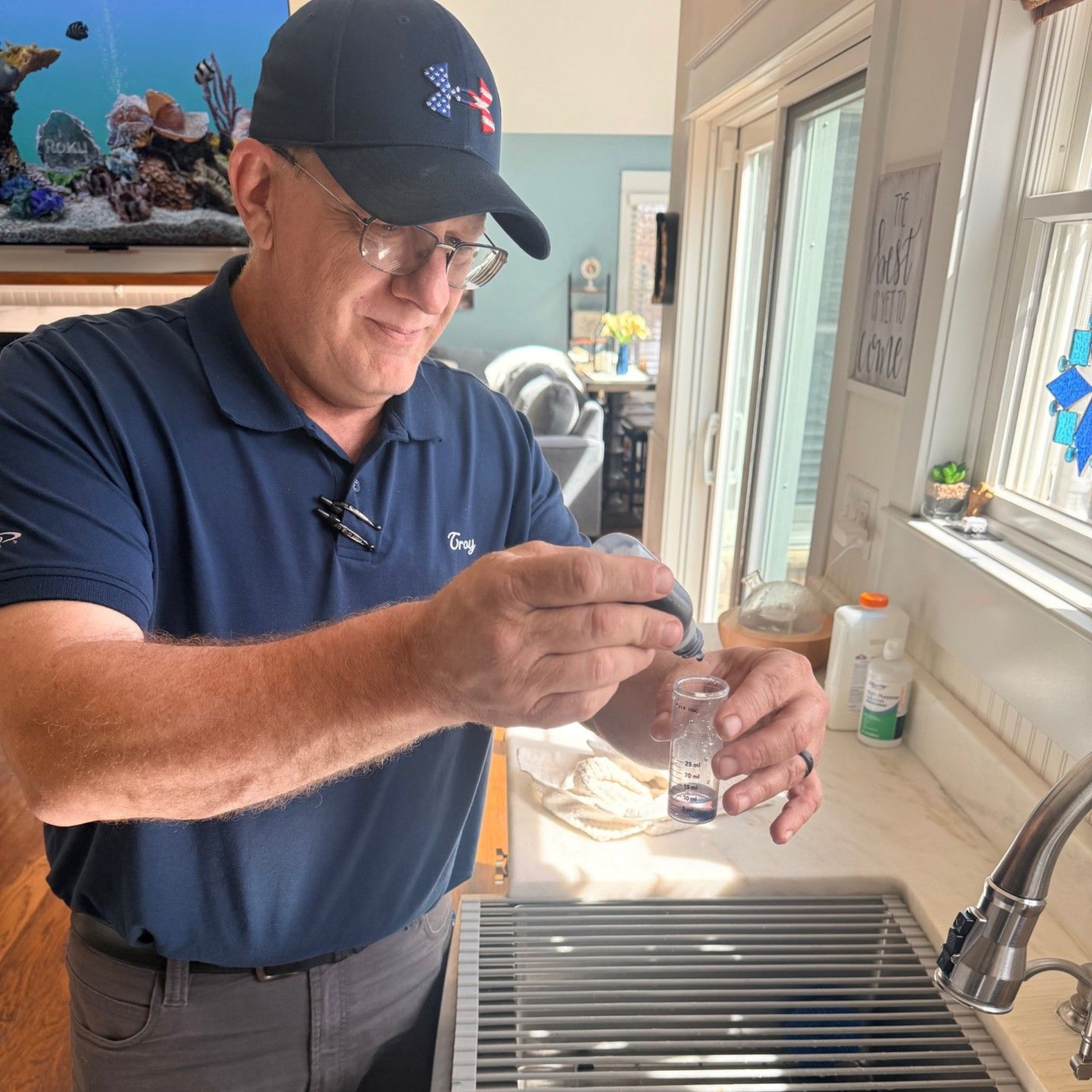 A person in a blue polo and cap adds drops of liquid from a small bottle into a test tube at a kitchen sink.