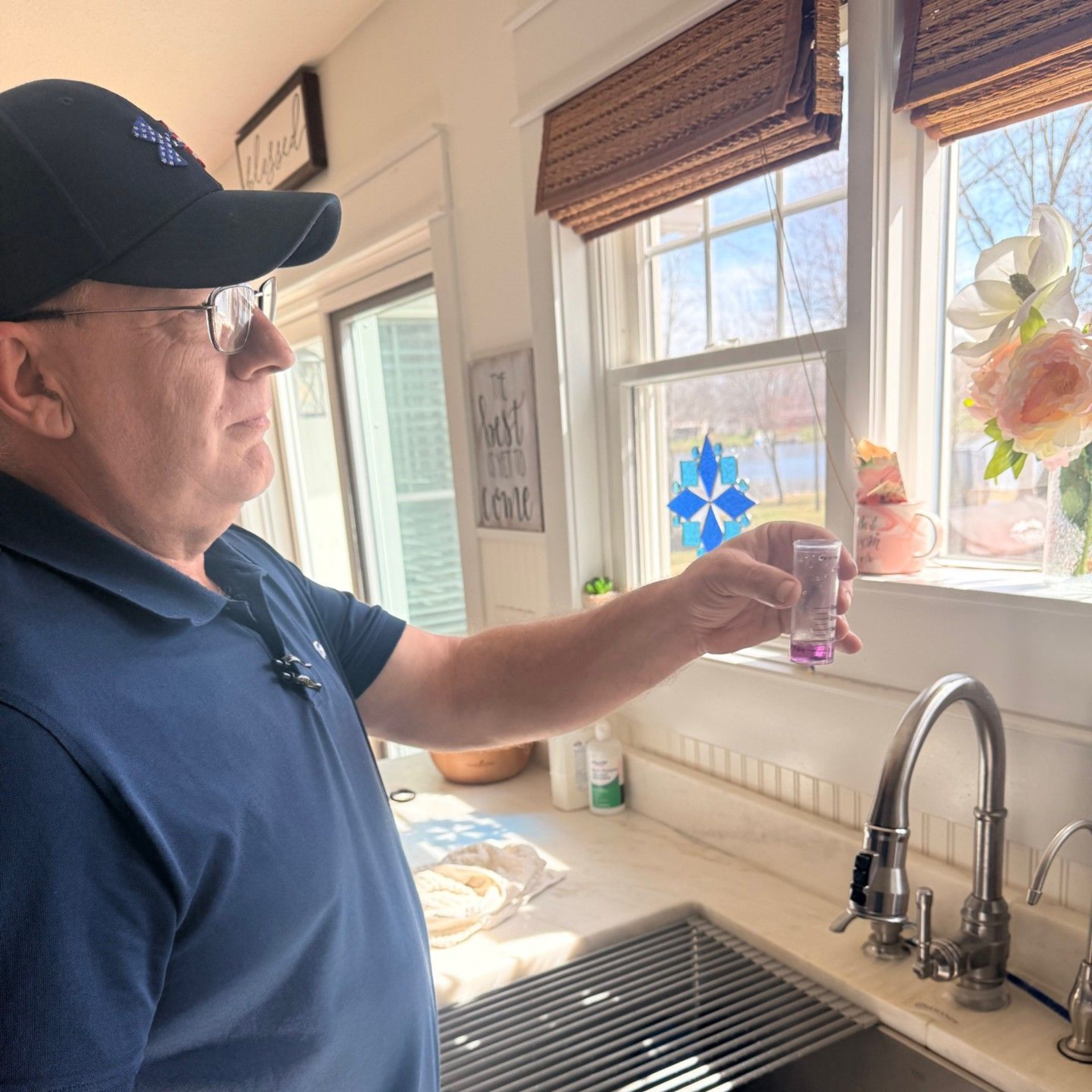 A person in a blue shirt standing at a kitchen sink, holding a test vial with purple liquid up to the window light.
