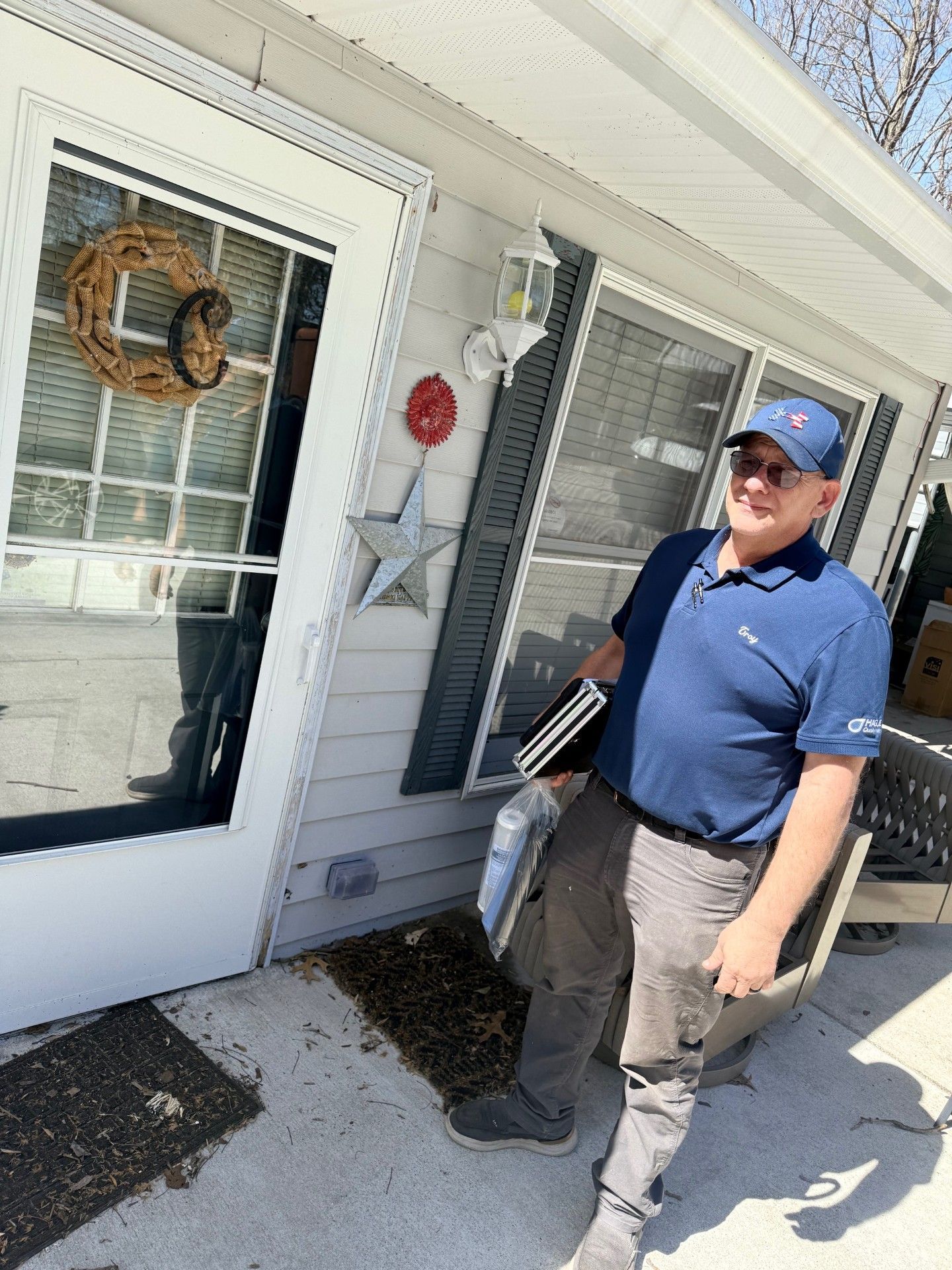 A smiling person in a blue polo and cap stands outside a home, holding a folder and supplies near a glass door.