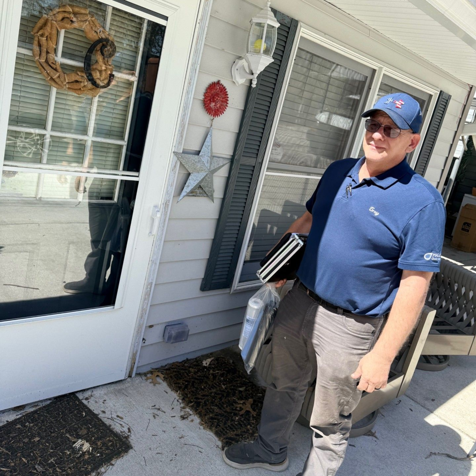 A smiling person in a blue polo and cap stands outside a home, holding a folder and supplies near a glass door.