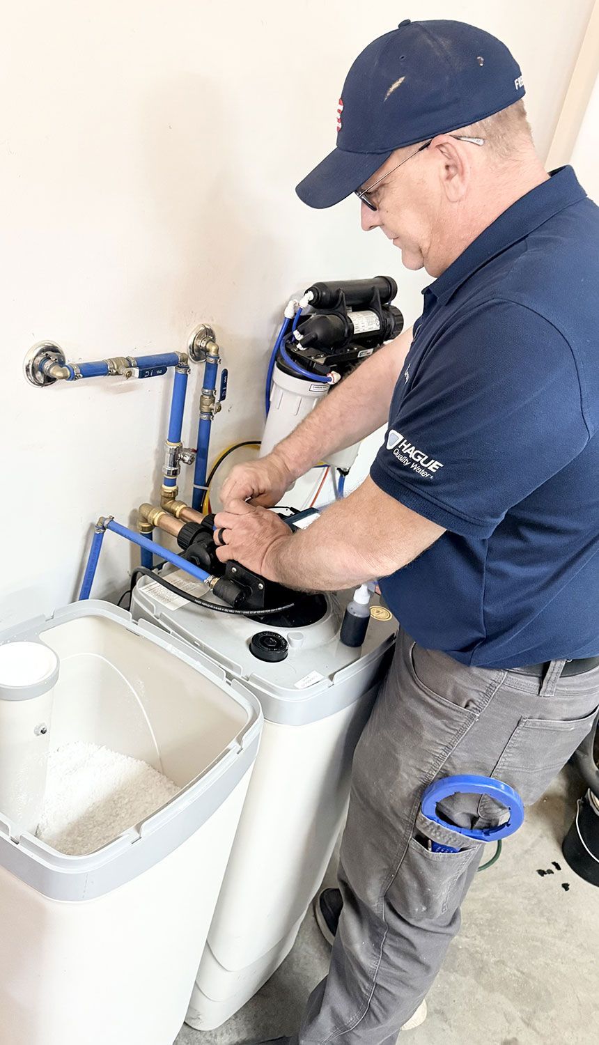 A technician in a blue uniform repairing a residential water softener system in a utility room.