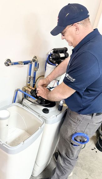 A technician in a blue uniform repairing a residential water softener system in a utility room.