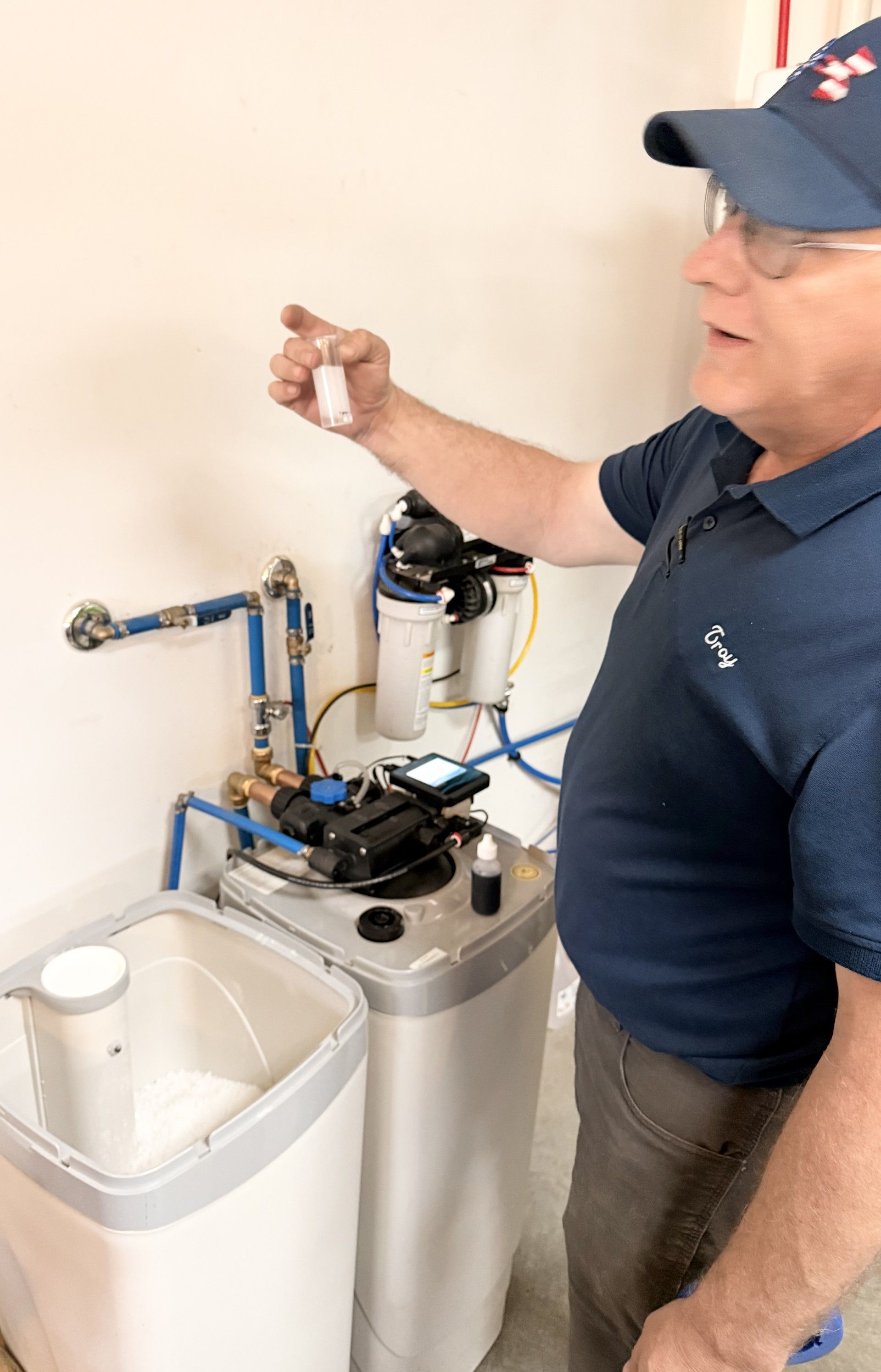 A person in a workshop stands next to a water softener system, holding up a small vial while explaining the equipment.