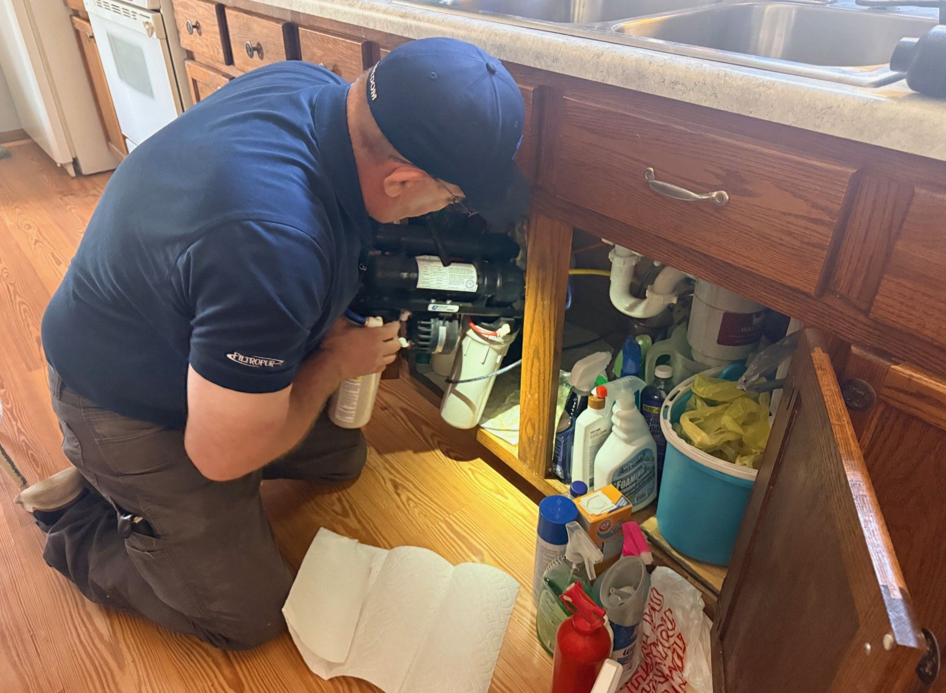 A person kneels under a kitchen sink to repair a water filtration system, surrounded by cleaning supplies and paper towels.