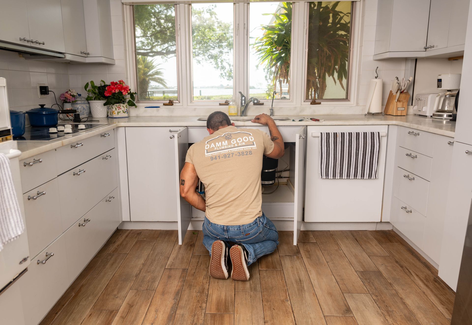 A person kneeling under a kitchen sink, working on plumbing. White cabinets, light wood floor, window in background.