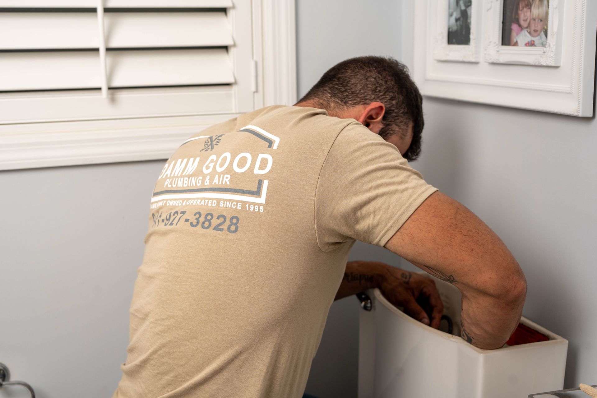 Plumber in a tan shirt works on a toilet tank in a bathroom.