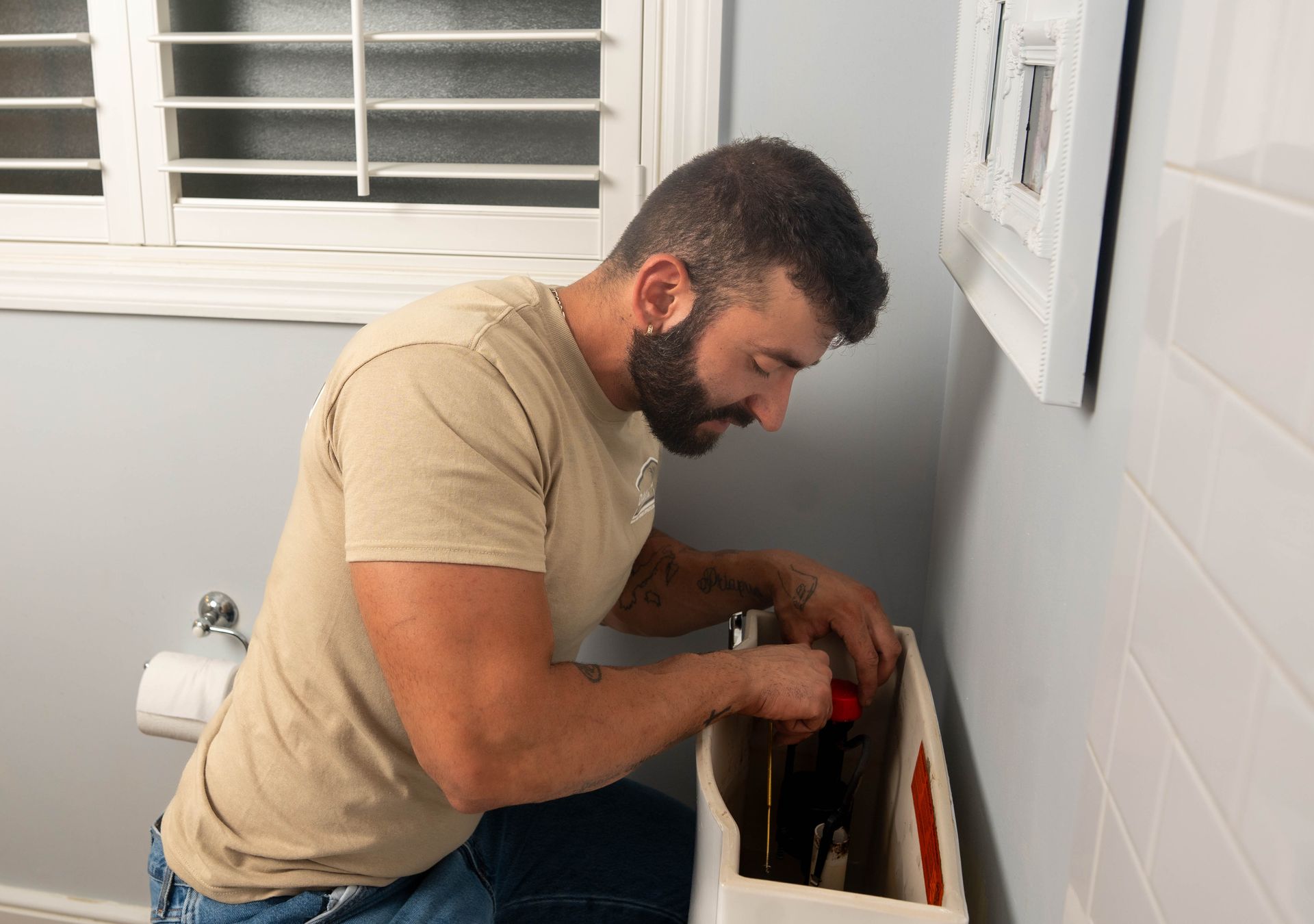 Man repairs toilet tank in bathroom. He is wearing a beige shirt and jeans, near a window and white wall.
