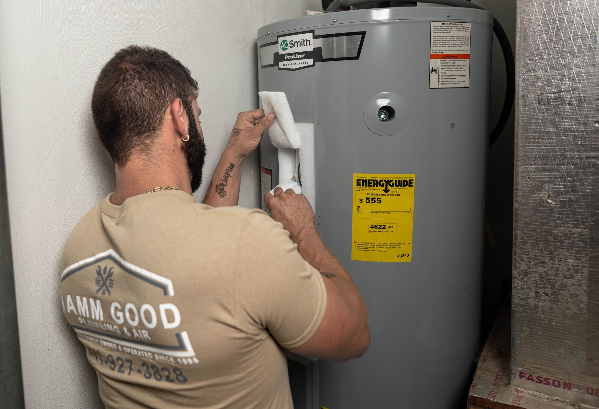 A person in a tan shirt installs insulation around a water heater in a utility room.