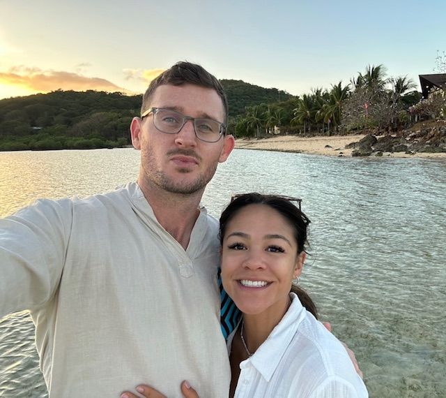 Couple smiling at the camera on a beach; water and greenery in the background.