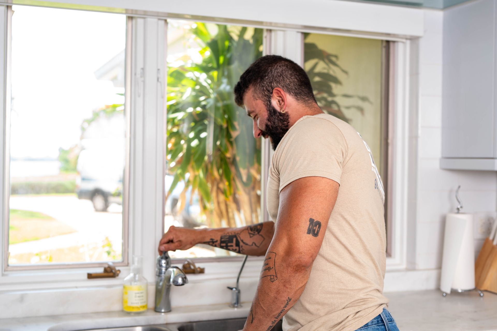 Man with arm tattoos at kitchen sink turning on faucet.