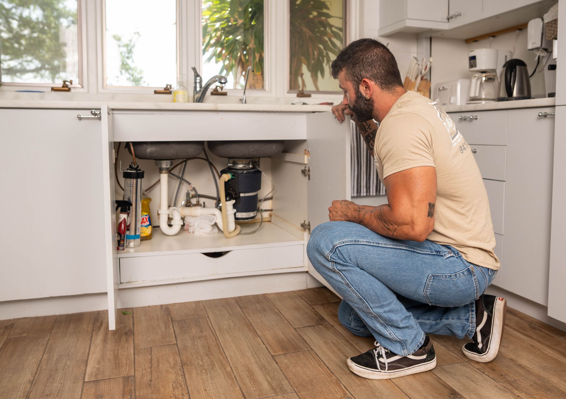 Man inspecting plumbing under a kitchen sink. He is crouching, looking at pipes and a disposal unit.