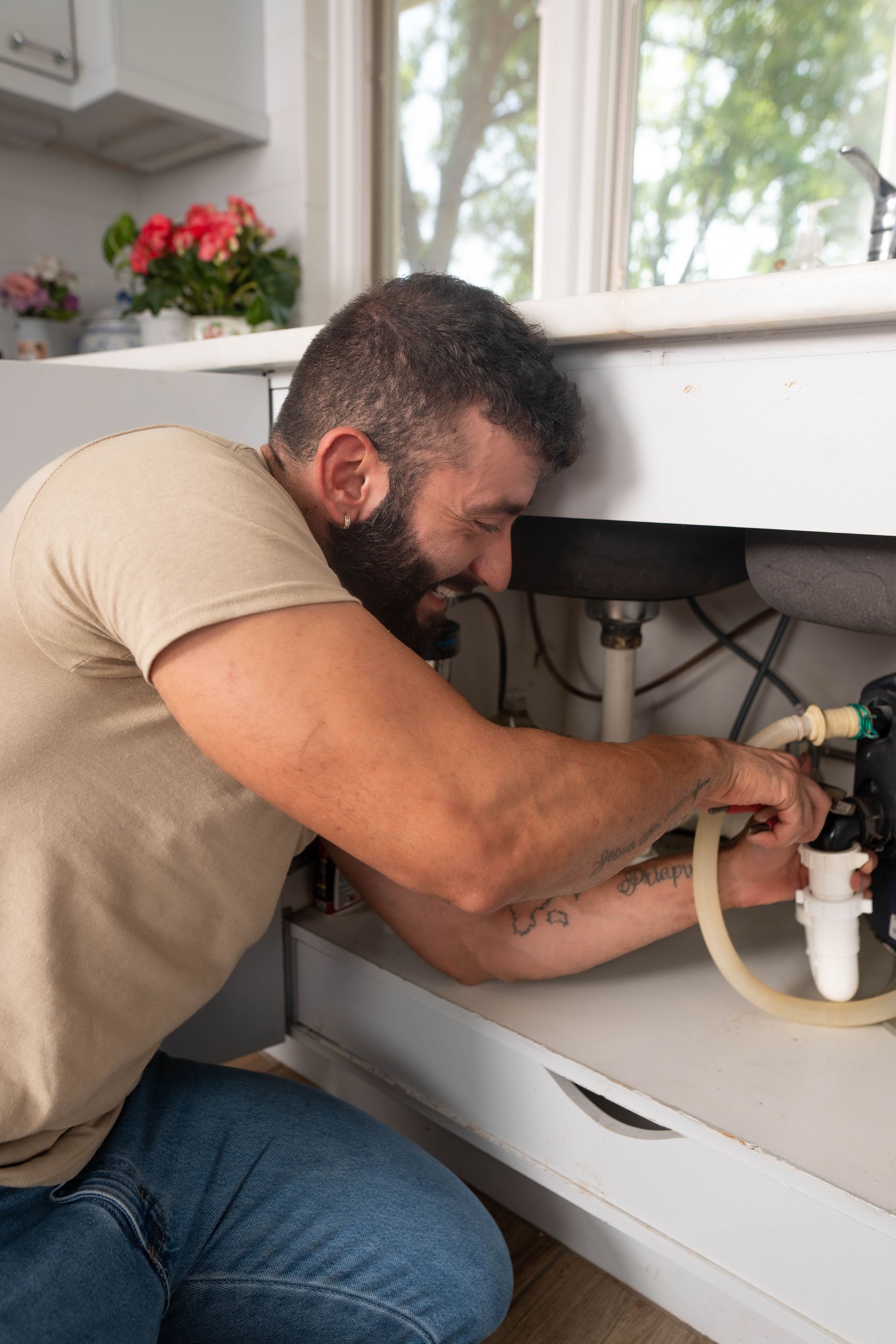 Man kneeling under a kitchen sink, fixing plumbing with a smile. Beige shirt, jeans. Interior setting.