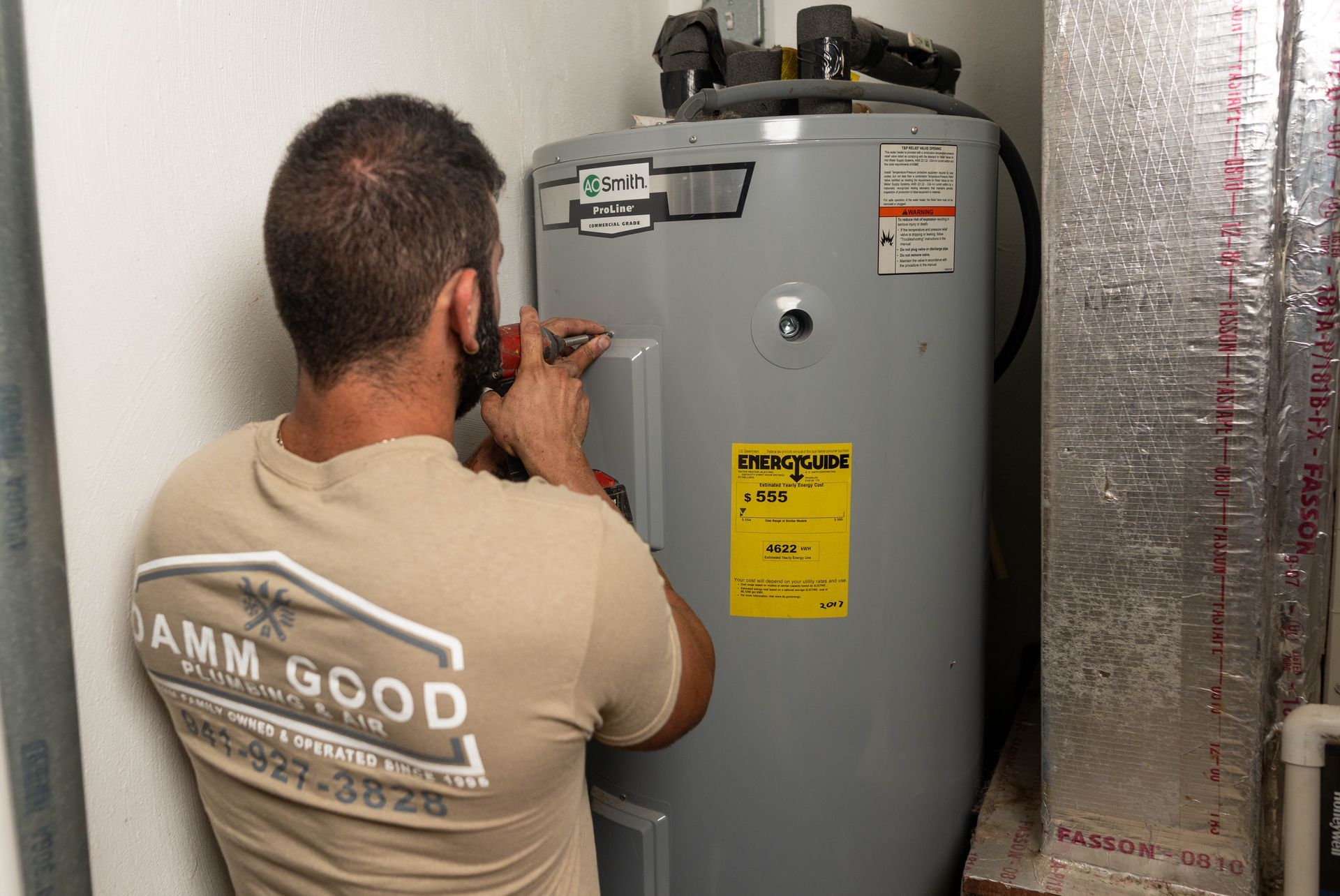 A man in a tan shirt works on a water heater, indoors. A 