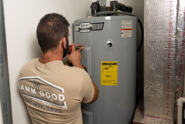 A person working on a water heater. The person is in a utility room and wearing a tan shirt with a logo.