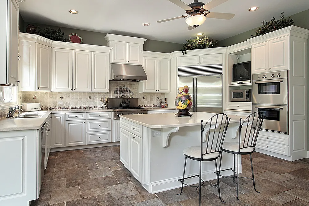 A kitchen with white cabinets , stainless steel appliances , a large island and a ceiling fan.